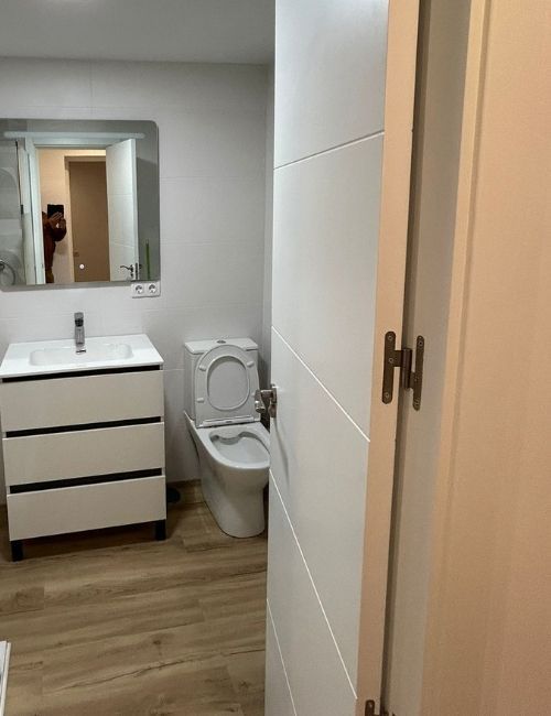 Baño con lavabo, inodoro y puerta blancos; suelo imitación madera. Se ve una persona en el espejo.