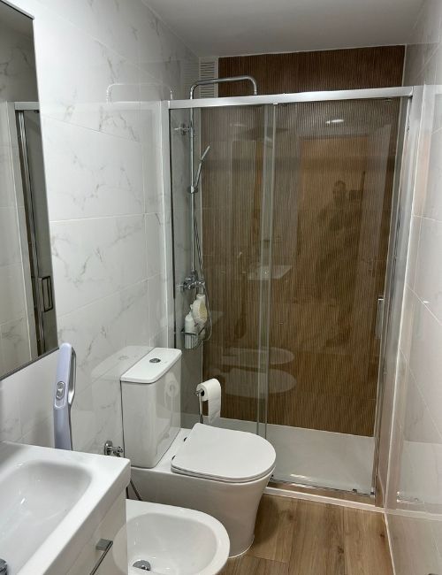Baño con inodoro blanco, bidé, lavabo y ducha de cristal. Paredes de mármol, ducha con respaldo de imitación madera y suelo imitación madera.