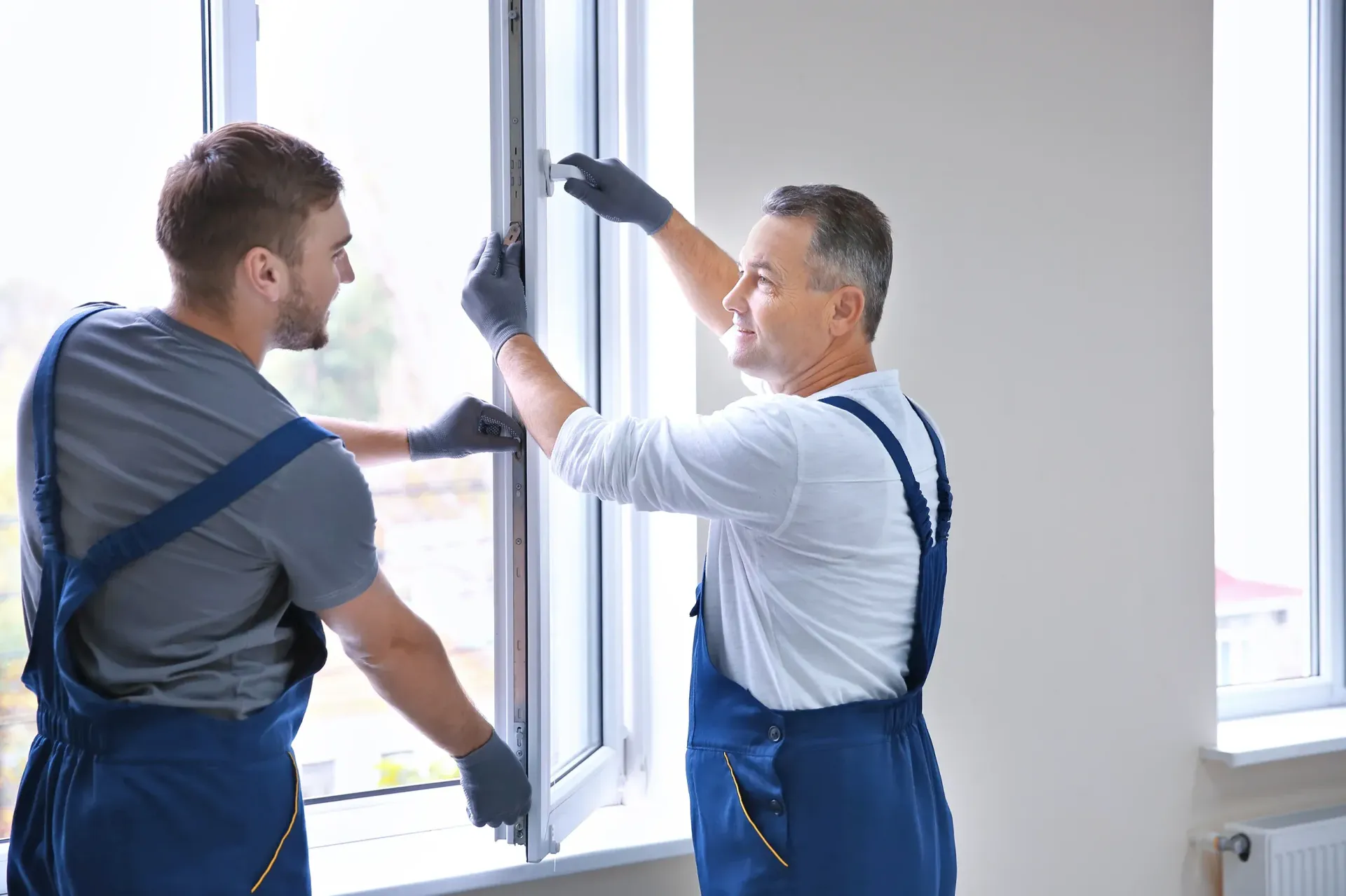 Dos hombres con overoles azules instalan una ventana; uno sonríe, el otro observa. Interior, paredes blancas.