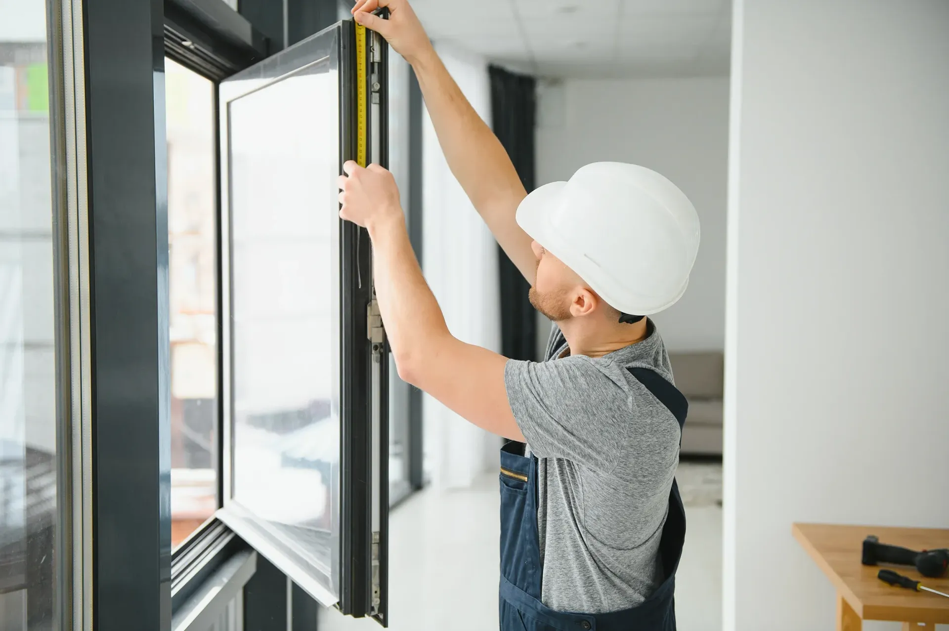 Un trabajador de la construcción con un casco blanco mide una ventana con una cinta métrica en una habitación luminosa.