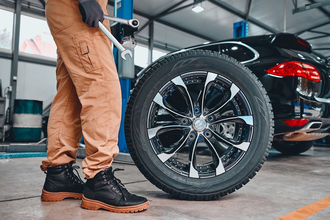 Un mécanicien en pantalon marron vérifie un gros pneu de voiture garé à côté d'une berline noire dans un garage.