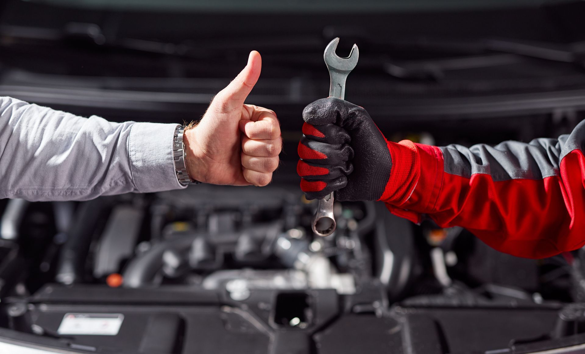 Deux mécaniciens, l'un ganté de rouge et l'autre de gris, échangent un signe d'approbation et une clé à molette dans un atelier de réparation automobile.