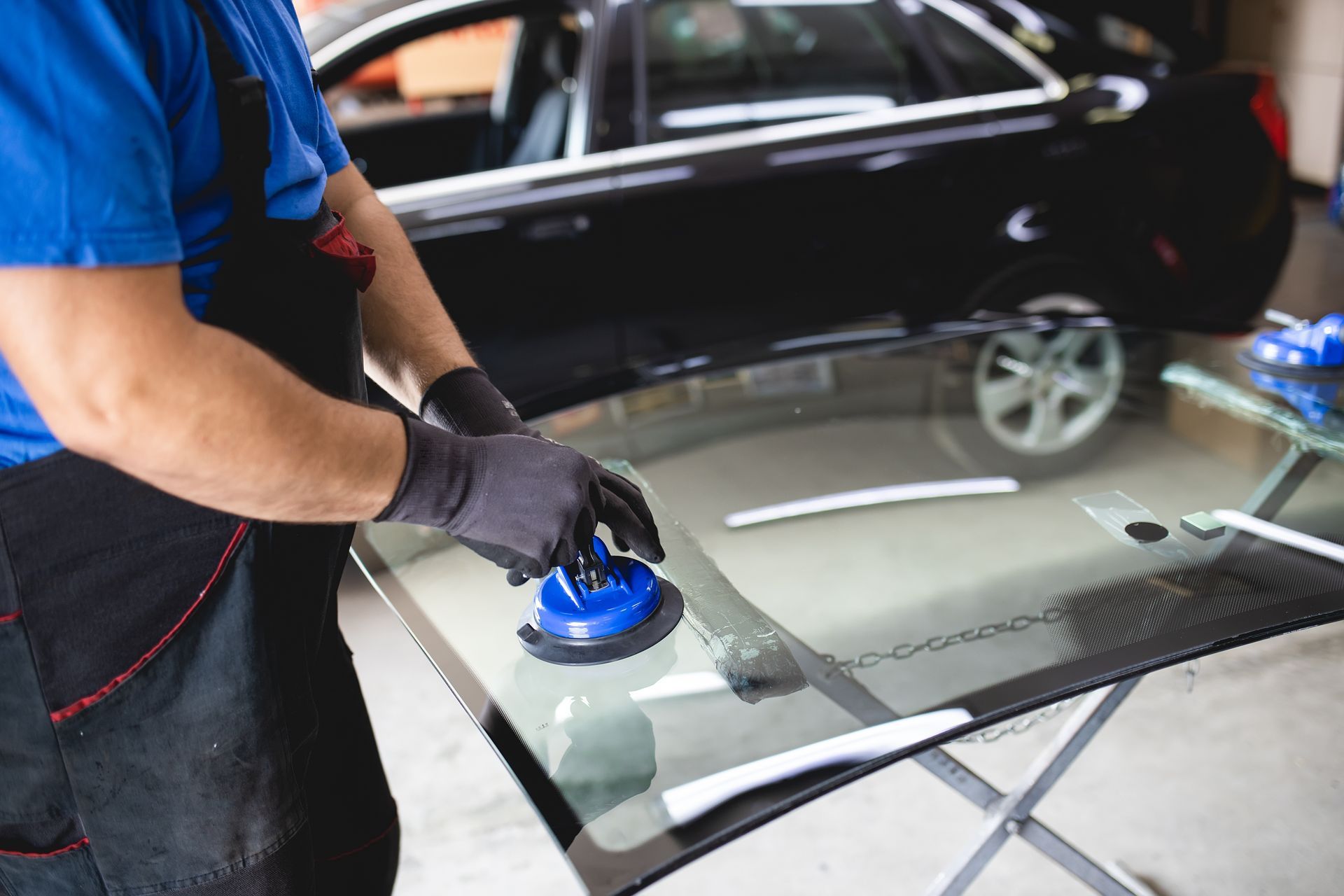 Un préparateur automobile polit le pare-brise d'une voiture avec un outil bleu dans un atelier de réparation automobile.