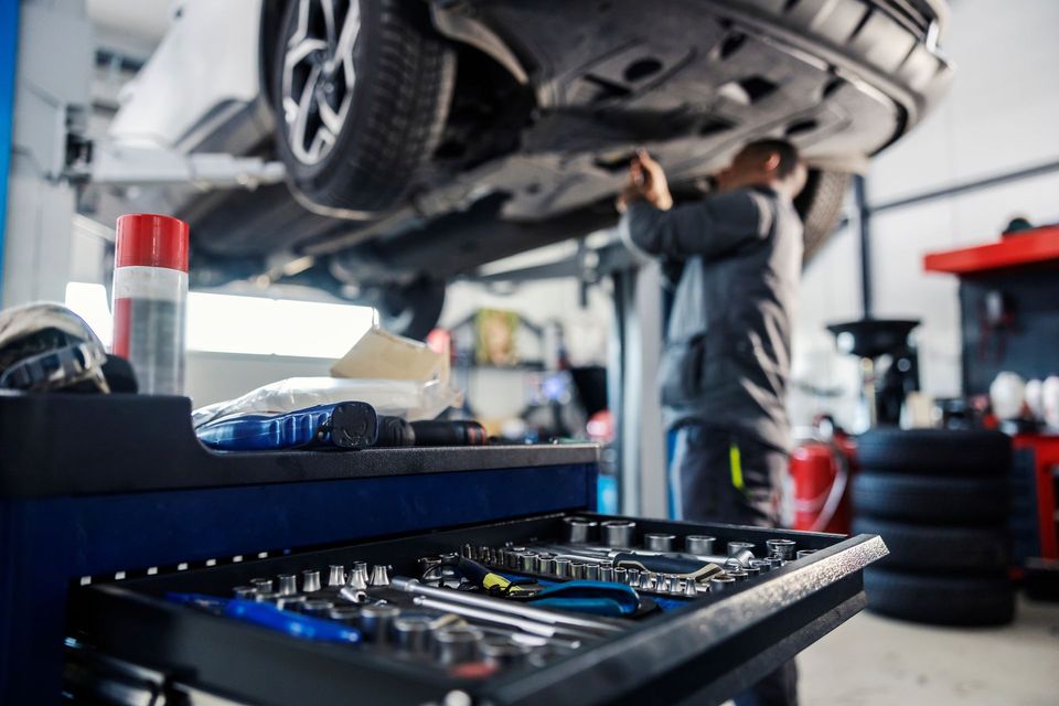 Un mécanicien travaille sous une voiture surélevée dans un atelier de réparation automobile, avec un plateau à outils au premier plan.