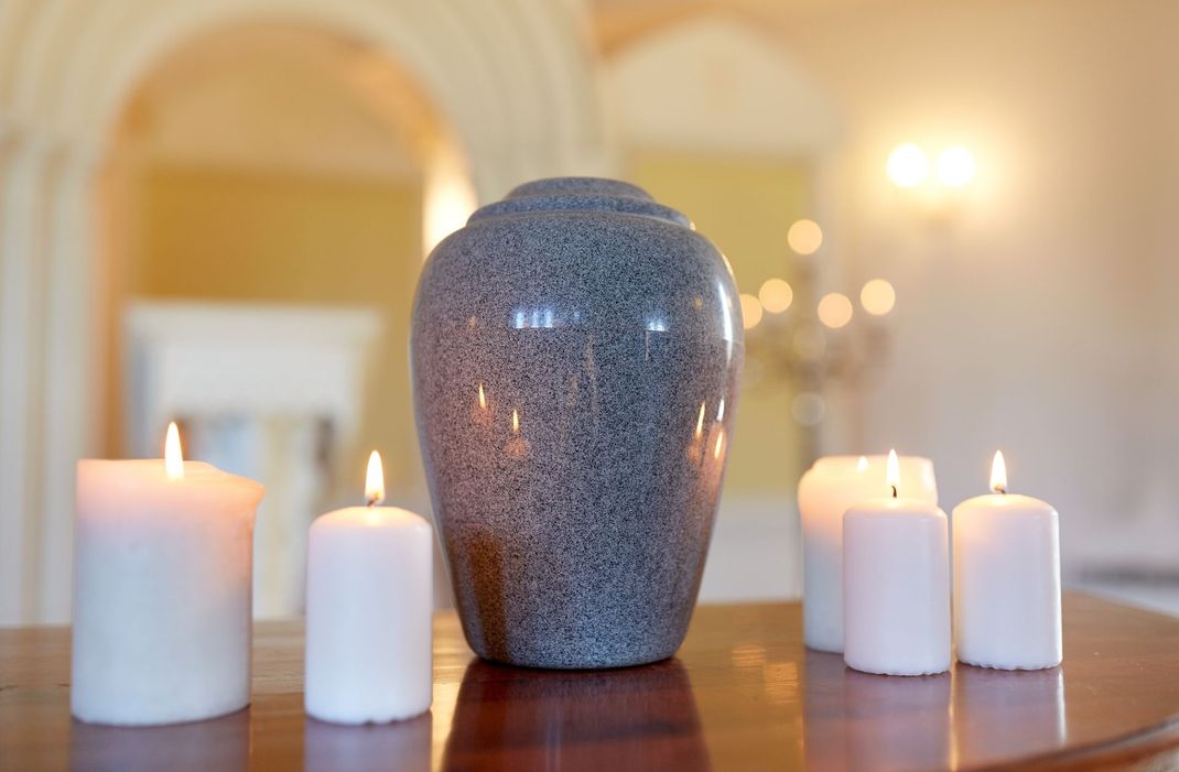 Cinq bougies blanches allumées entourent une urne bleu-gris sur une table cirée dans une pièce à l'éclairage tamisé.