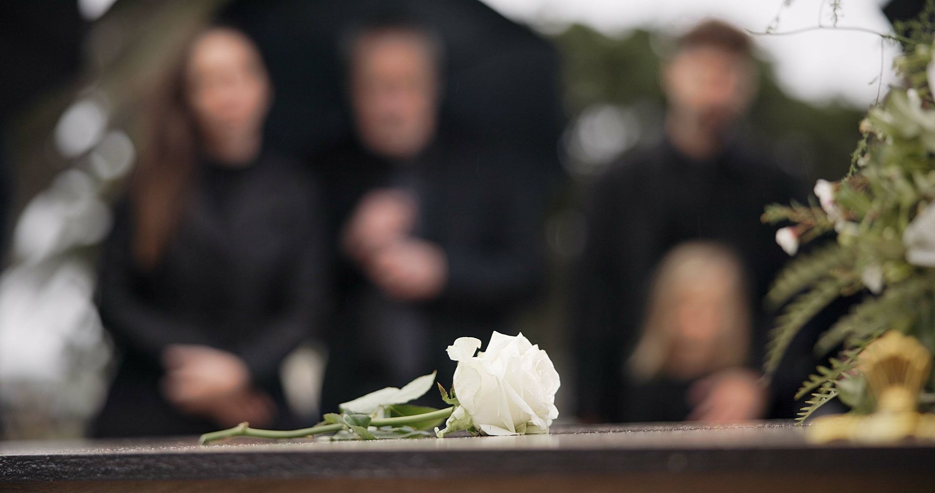 Une rose blanche sur un cercueil en bois, avec des personnes en deuil floues en arrière-plan.
