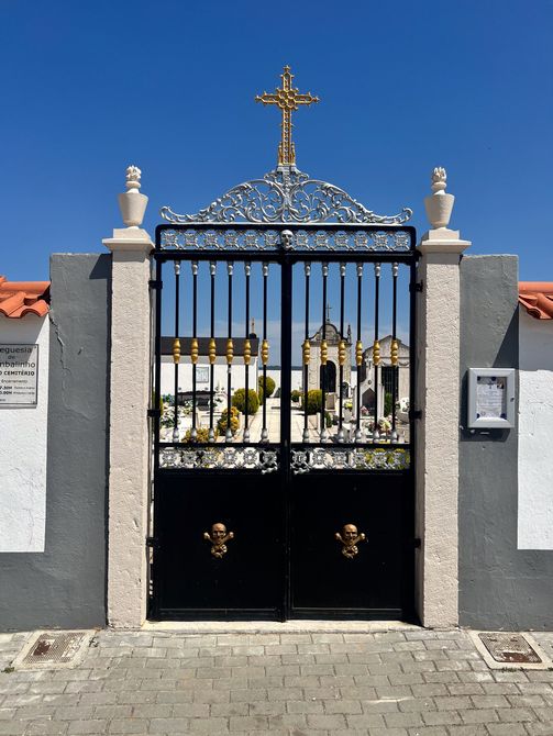 Portail noir orné d'une croix dorée et de piliers en pierre blanche à l'entrée d'un cimetière ensoleillé.