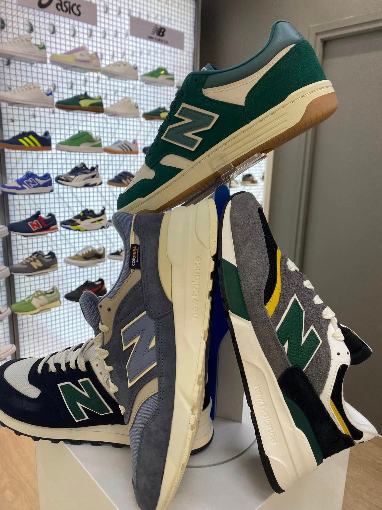 Pile de baskets New Balance vertes, bleu marine et crème sur un mur d'exposition dans un magasin de chaussures