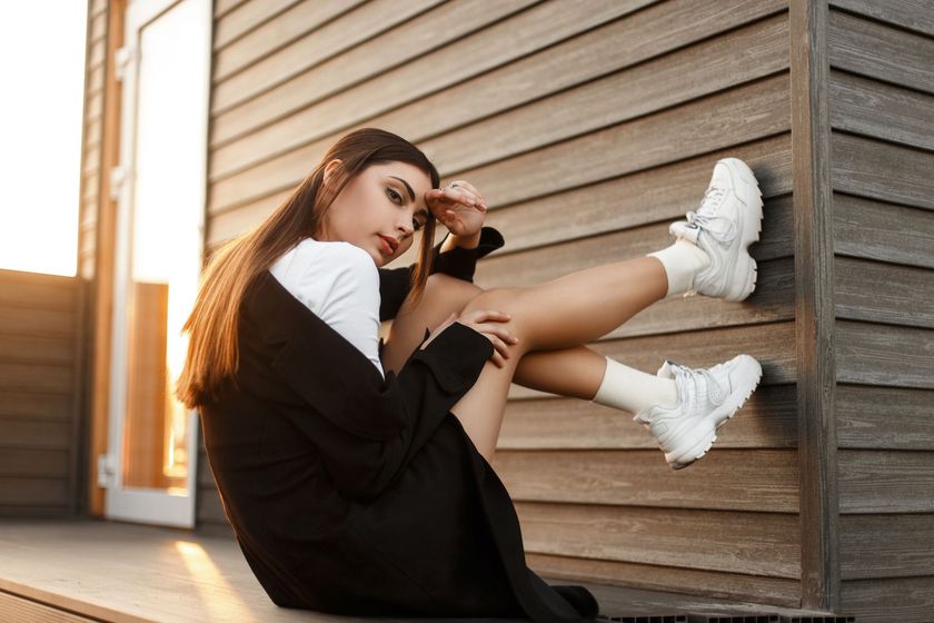 Jeune femme assise contre un mur en bois, les genoux repliés contre sa poitrine, baignée par la douce lumière du soleil, vêtue de noir et blanc et de baskets blanches.