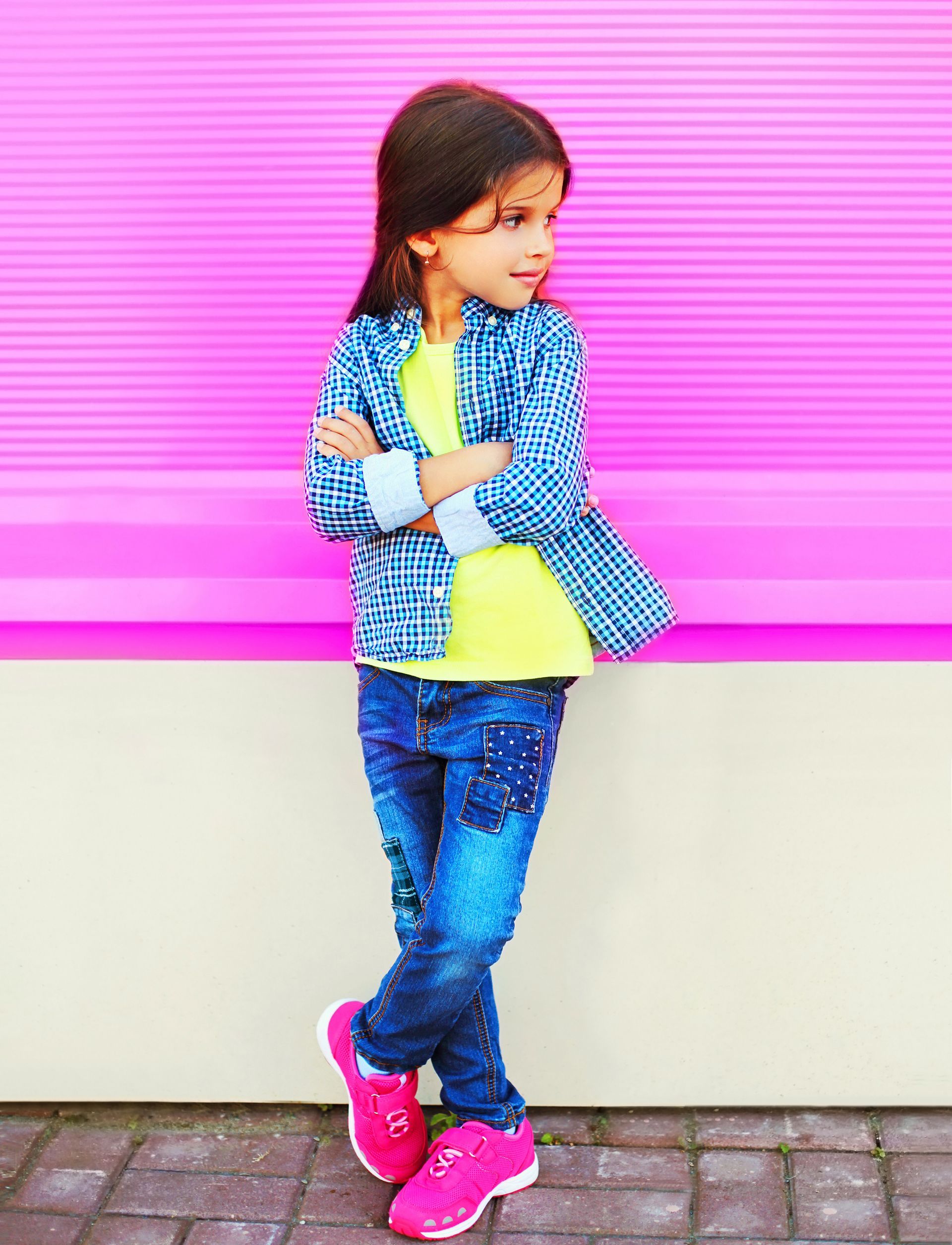 Enfant en veste bleue et jean appuyé contre un mur rose, portant des baskets rose vif