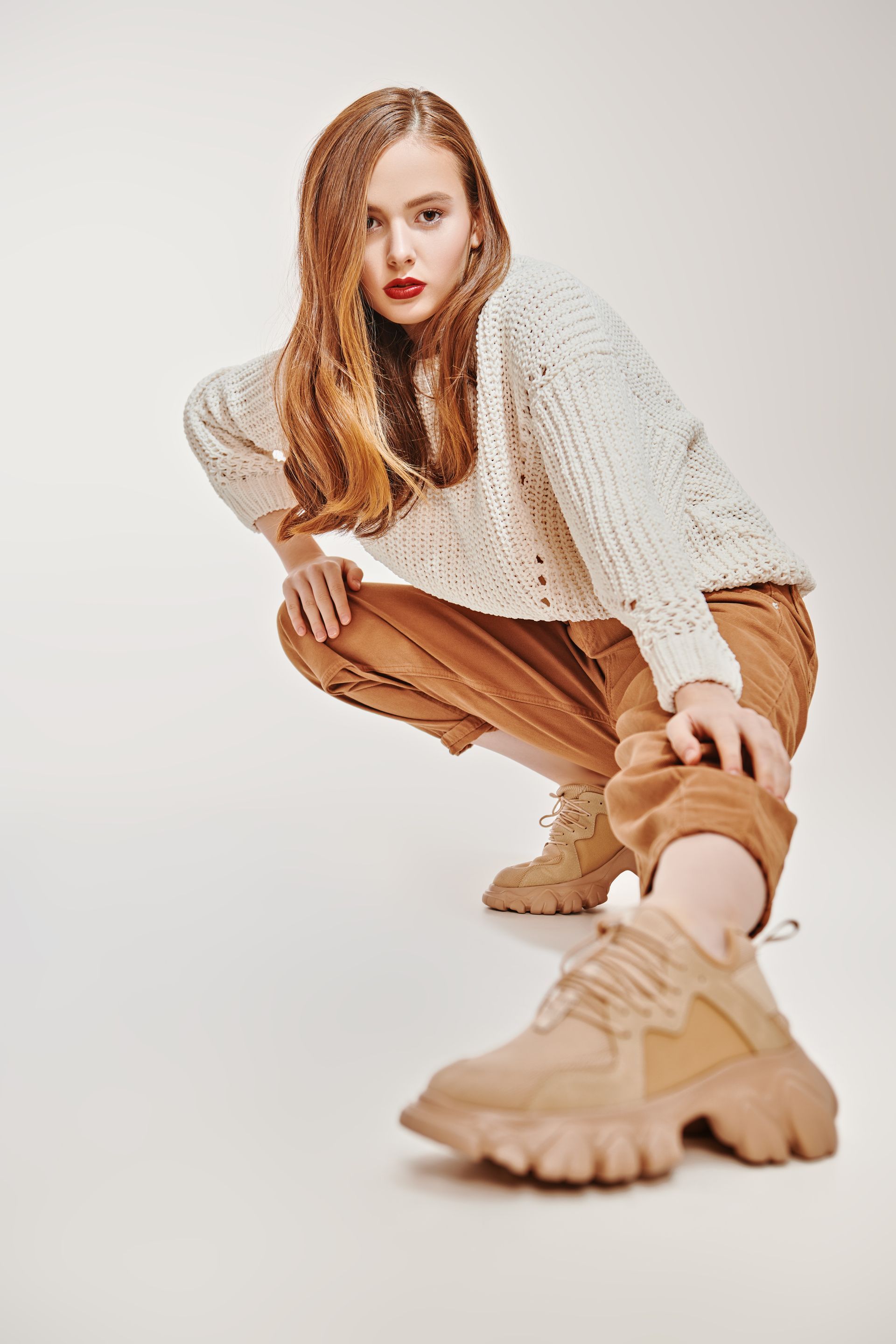 Mannequin accroupie, vêtue d'un pull en maille beige, d'un pantalon camel et de bottines à semelles épaisses, sur fond blanc studio.
