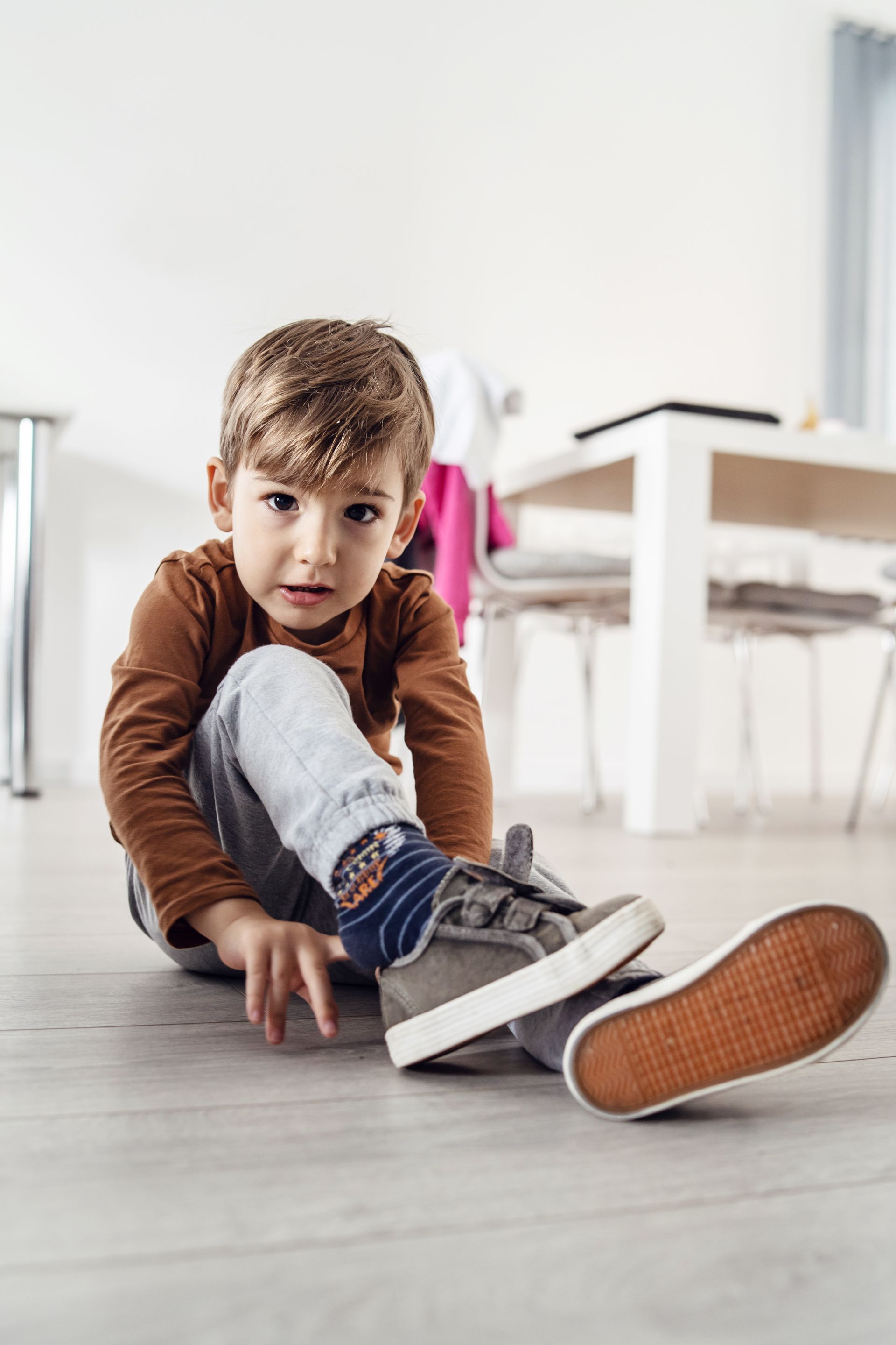 Un enfant noue une basket grise assis sur un parquet clair dans une pièce lumineuse