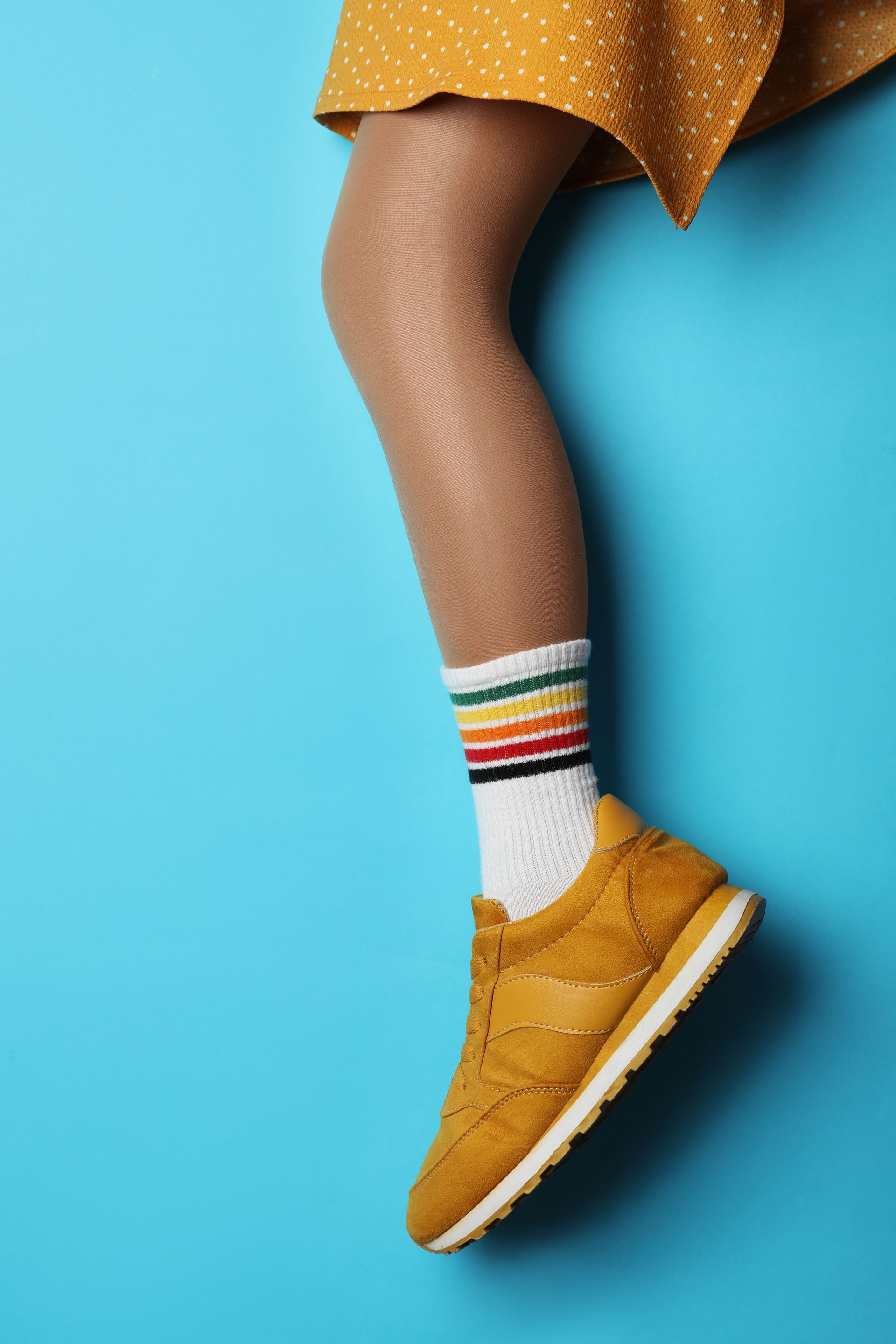 Jambes vêtues d'une robe moutarde, de chaussettes blanches à rayures et de baskets moutarde sur fond bleu