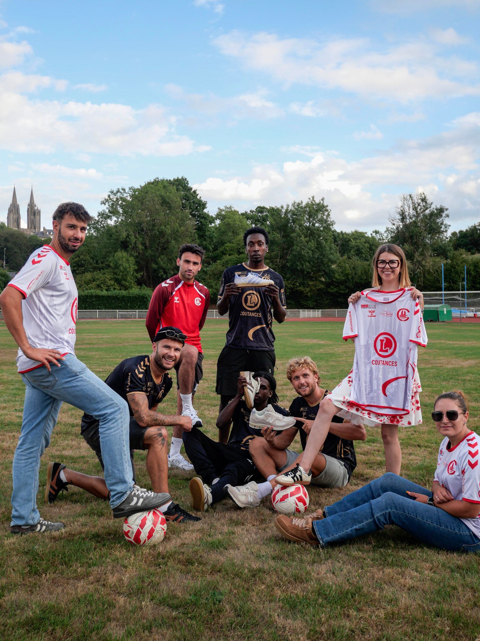 Un groupe pose sur un terrain herbeux avec des ballons de football, portant des maillots et des vêtements décontractés sous un ciel bleu.