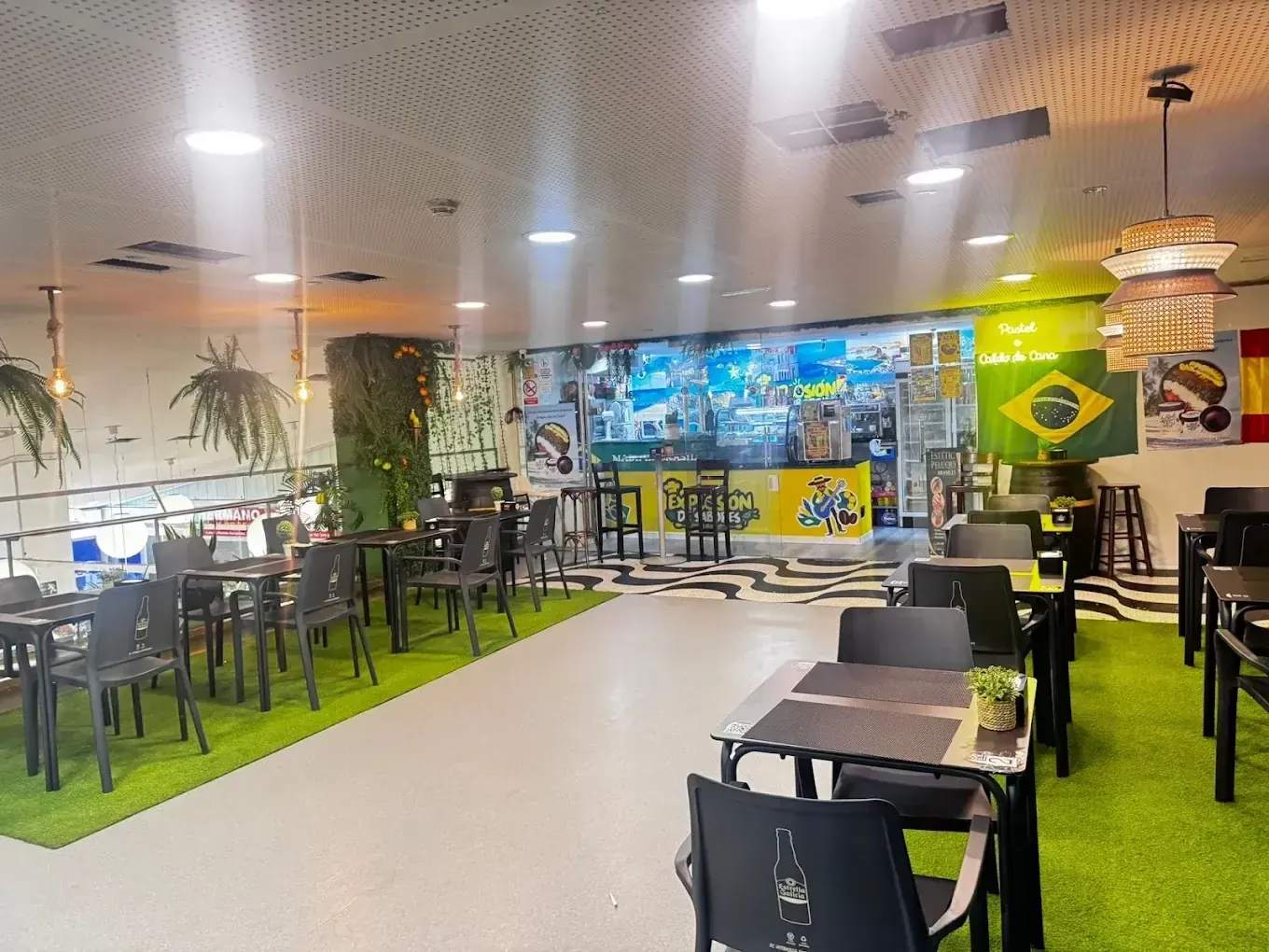 Interior de un restaurante bien iluminado con mesas, sillas y zona de bar, con decoración brasileña y española.