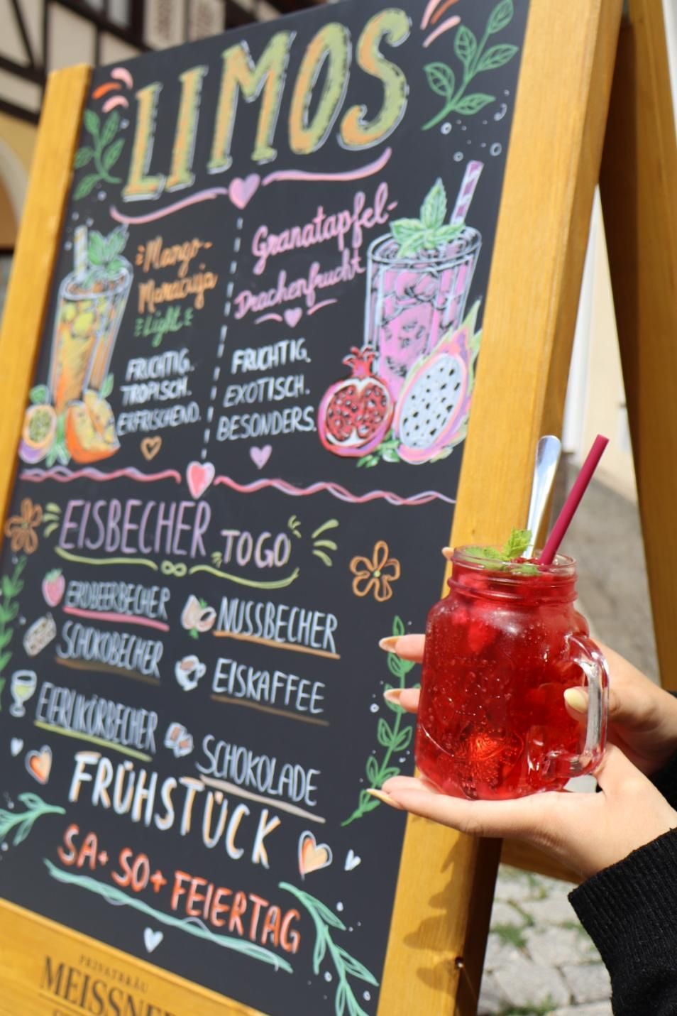 Kreidetafel-Café-Schild, das Limousinen und Desserts anpreist, daneben eine Hand, die ein rotes Eisgetränk hält.