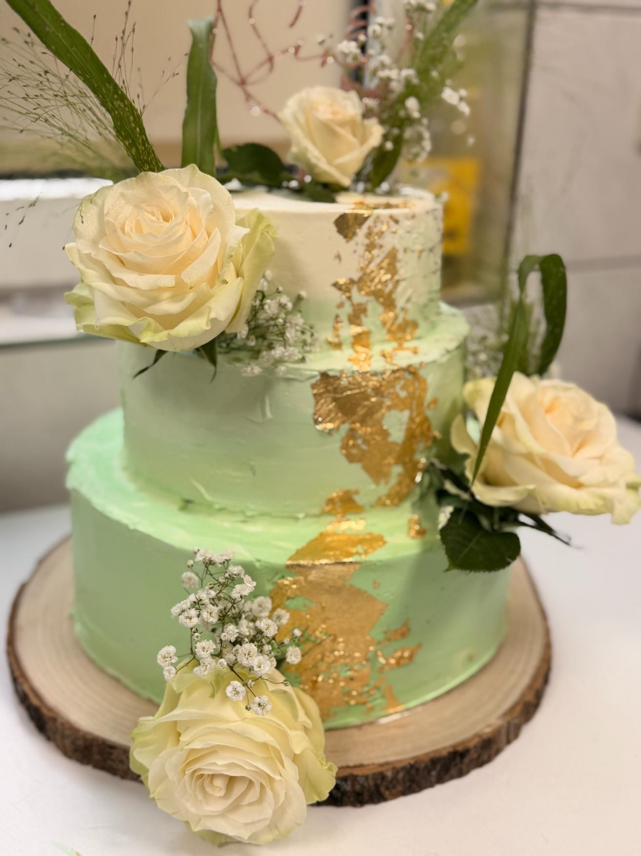 Zweistöckige mintgrüne Torte mit cremefarbenen Rosen, goldenen Akzenten und Grünzeug auf einem Holzsockel