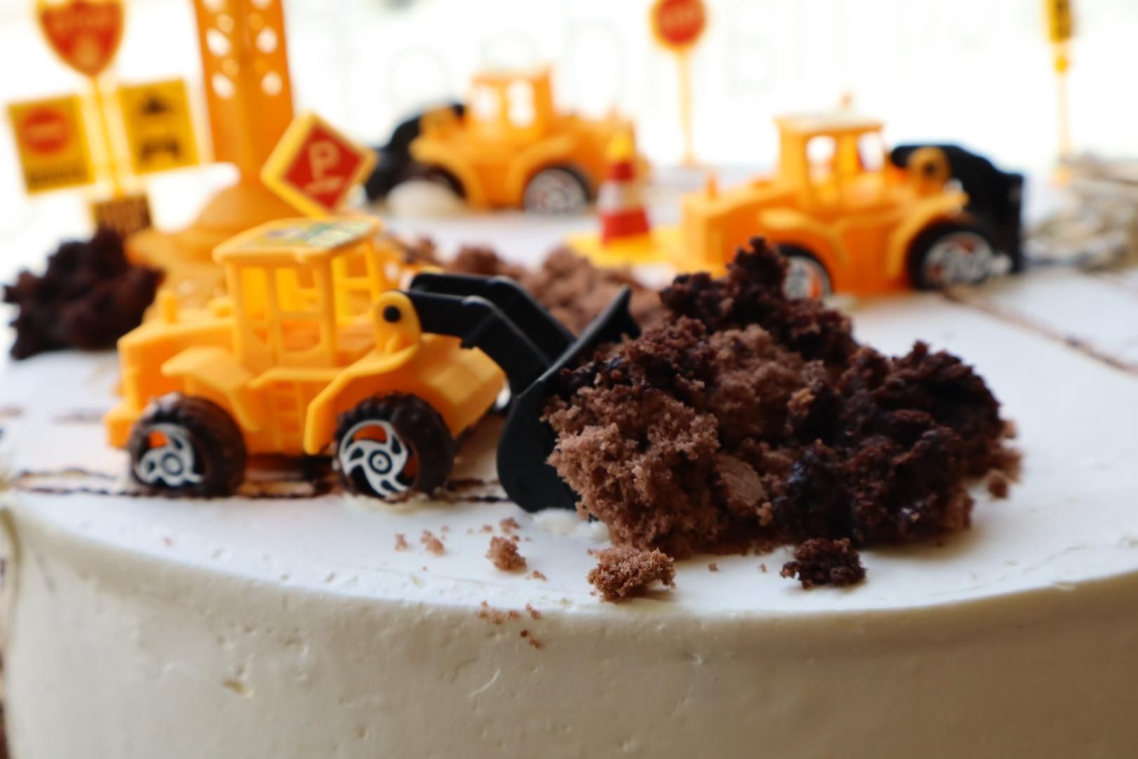 Ein Spielzeugbulldozer schiebt Schokoladenkrümel auf einen weiß glasierten Kuchen mit Baustellenfahrzeugen.