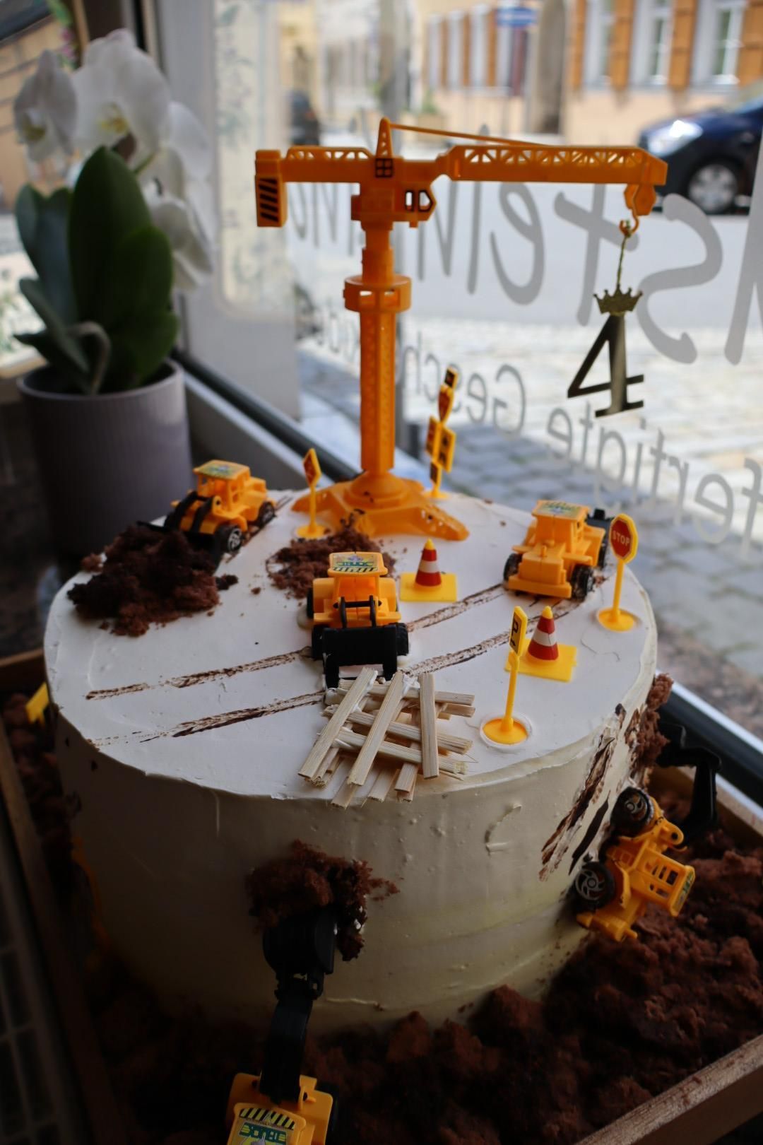Gelber Spielzeugkran auf einer weißen Torte mit Mini-Baufahrzeugen vor einem Caféfenster