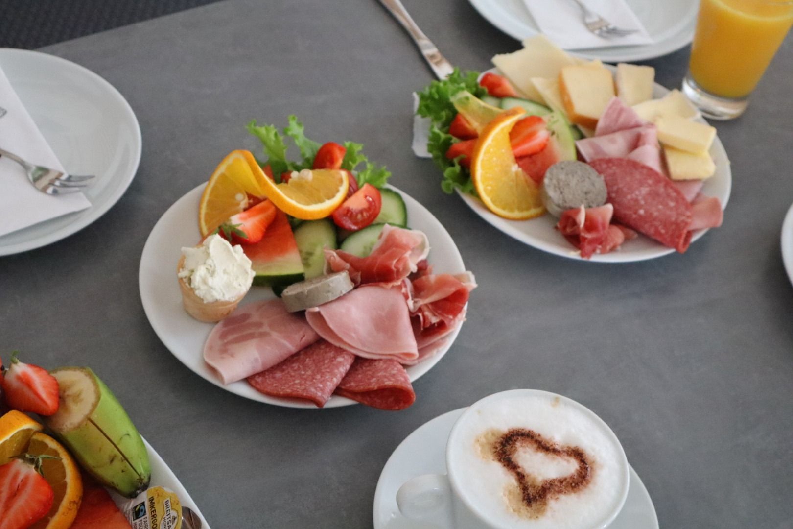 Teller mit verschiedenen Aufschnittsorten, Käse, Obst und Salat auf einem Esstisch mit Kaffee