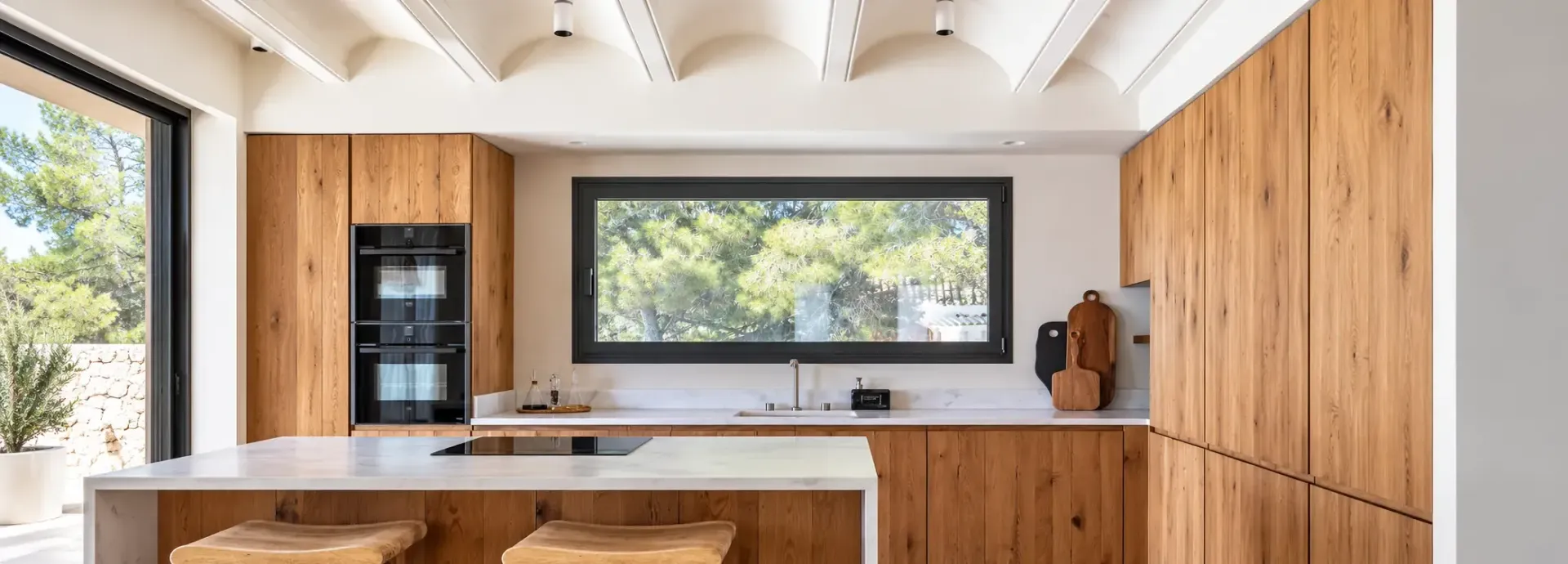 Cocina moderna con armarios de madera, isla blanca, electrodomésticos negros y grandes ventanales con vistas al exterior
