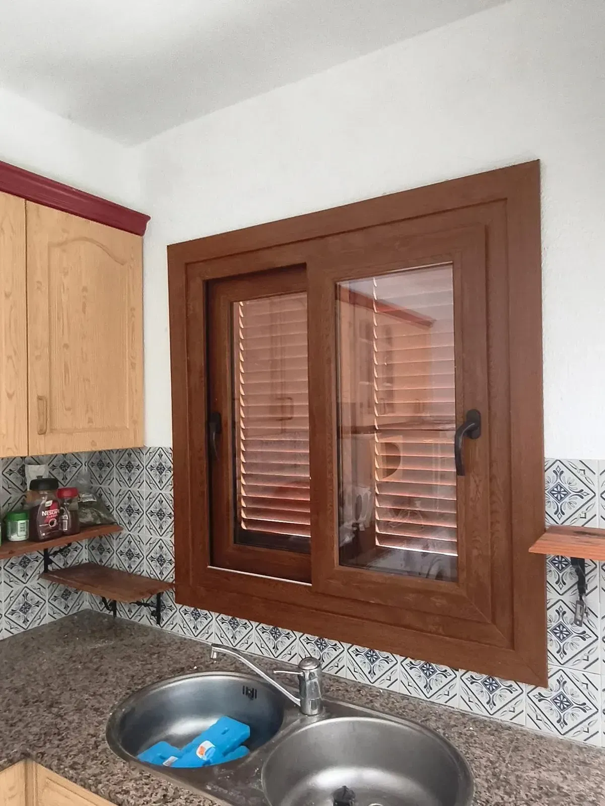 Fregadero de cocina bajo una ventana con marco marrón, contraventanas de madera y un salpicadero de azulejos estampados.