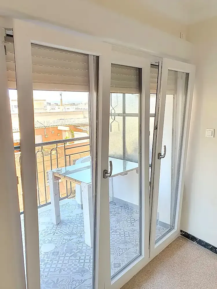 Las puertas de cristal del balcón se abren a una luminosa terraza al aire libre con una mesa y sillas.