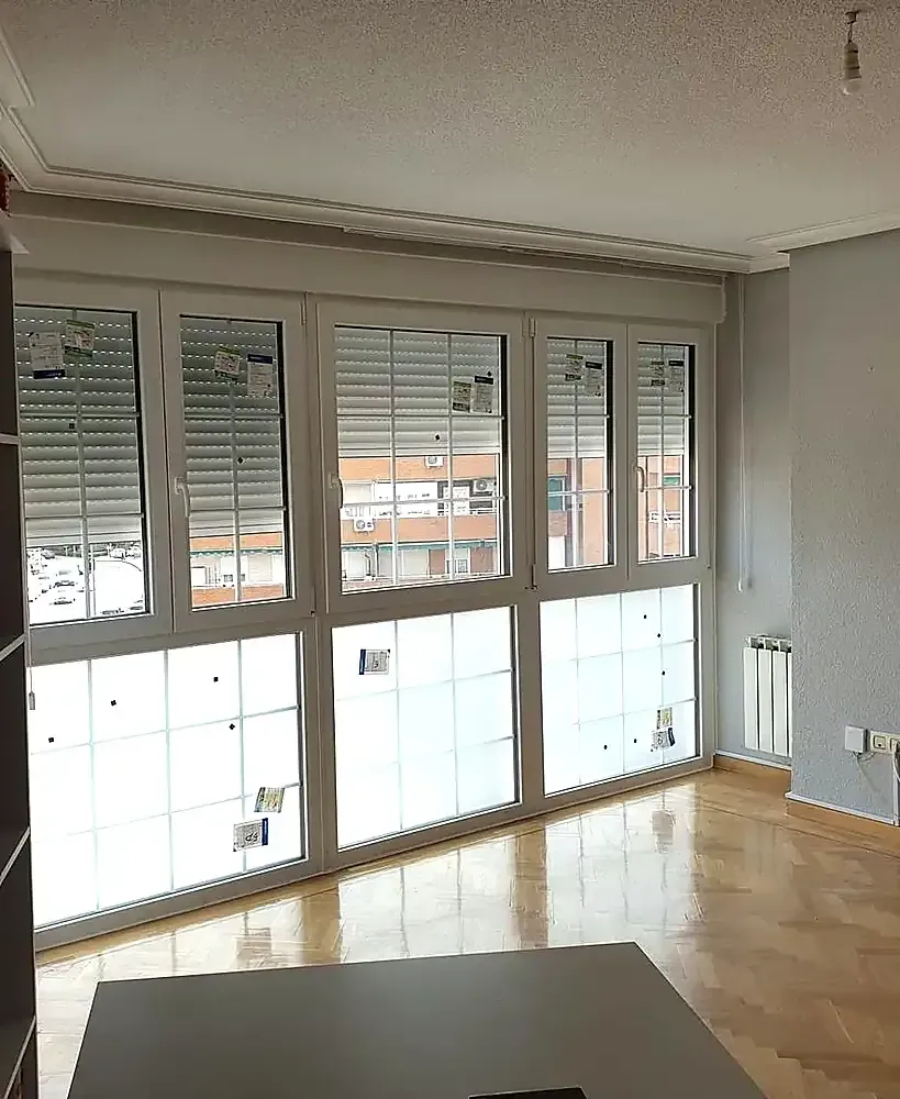 Salón vacío con suelo de parquet y grandes ventanales con persianas blancas.