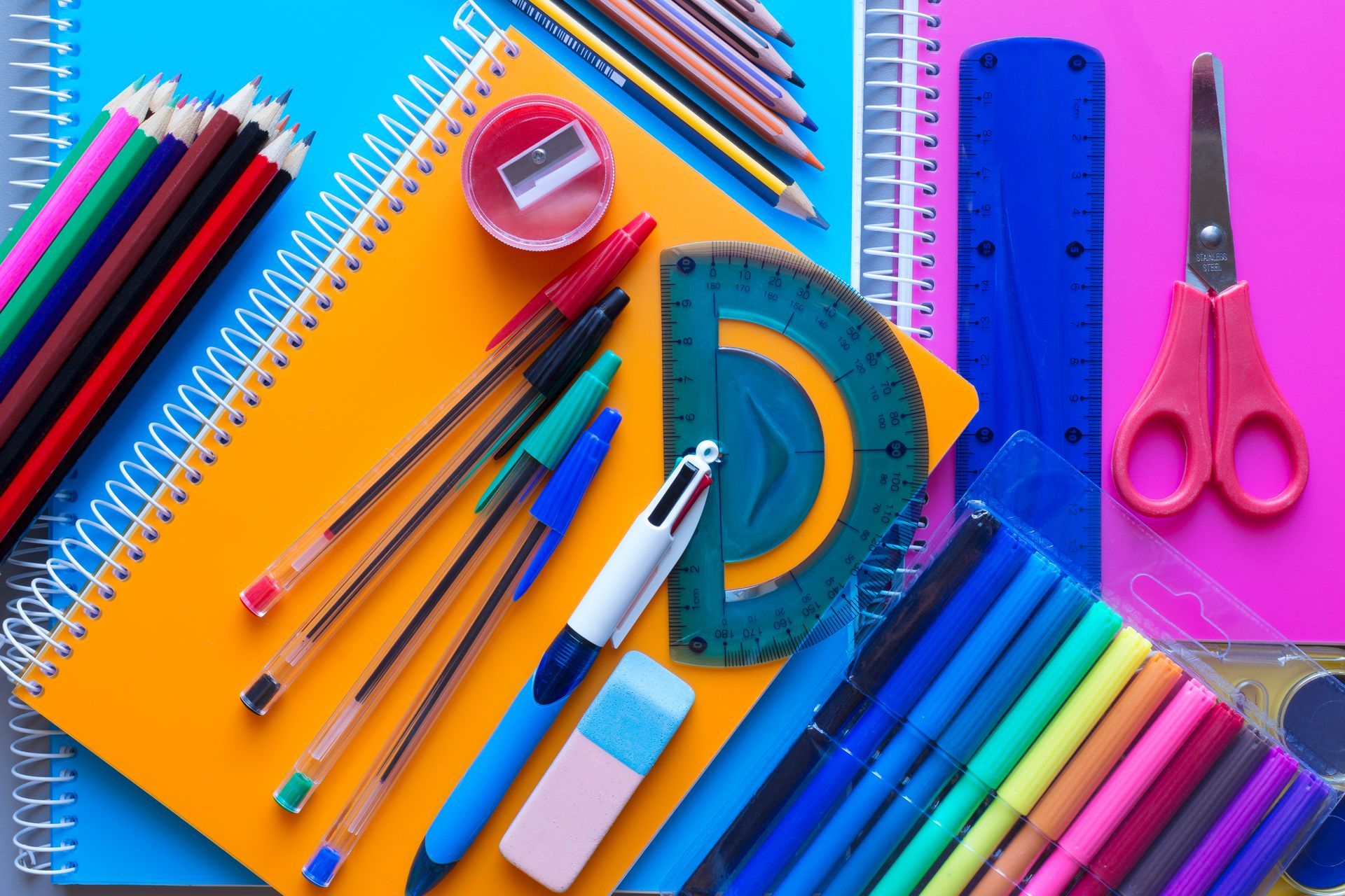 Un assortiment coloré de fournitures scolaires, comprenant des cahiers, des stylos, des ciseaux et un rapporteur.