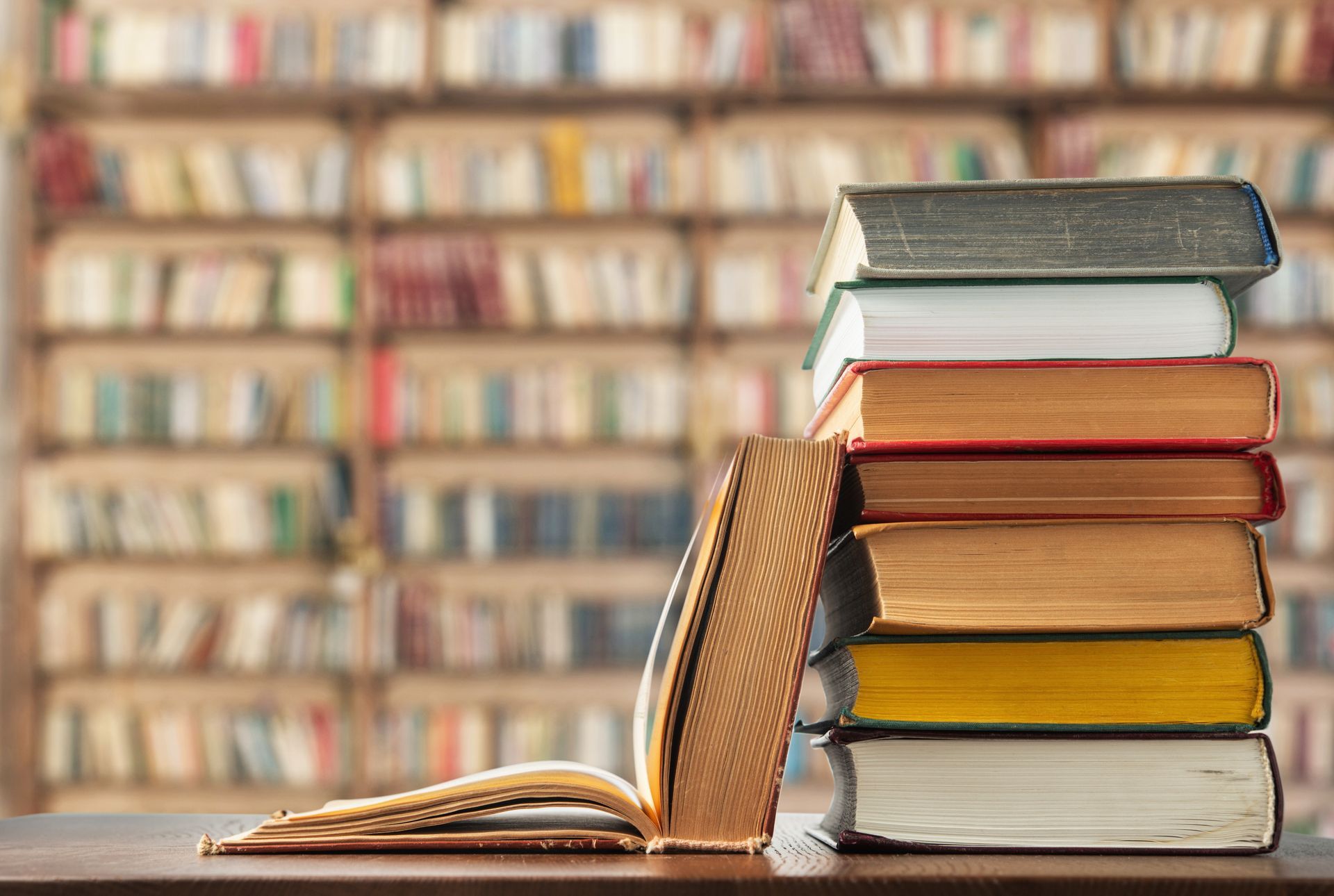 Une pile de livres multicolores est posée à côté d'un livre ancien ouvert, devant une étagère de bibliothèque.