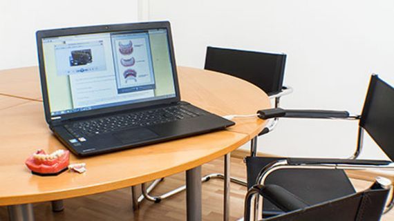 Auf einem Tisch steht ein Laptop mit Zahndiagrammen; Modellzähne und Stühle befinden sich in der Nähe.