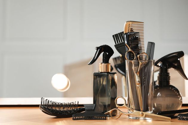 Une coiffeuse coupe les cheveux d'une cliente assise dans un salon, avec un miroir et des plantes à proximité.