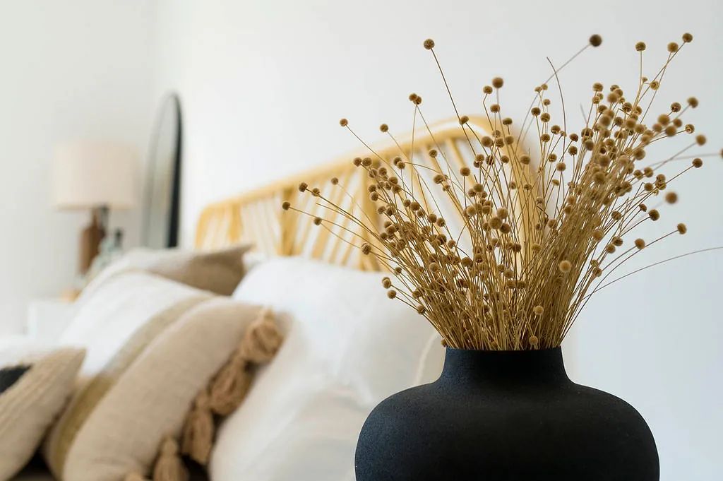 Un jarrón negro lleno de flores secas está frente a una cama.