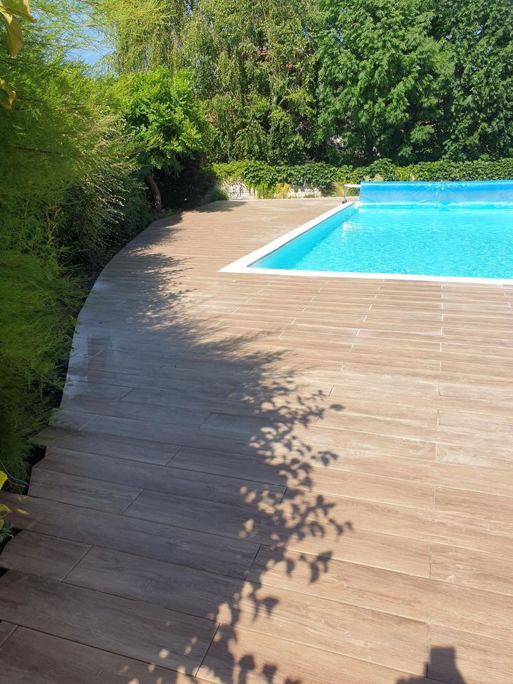 Terrasse en bois incurvée autour d'une piscine bleue, entourée de verdure.