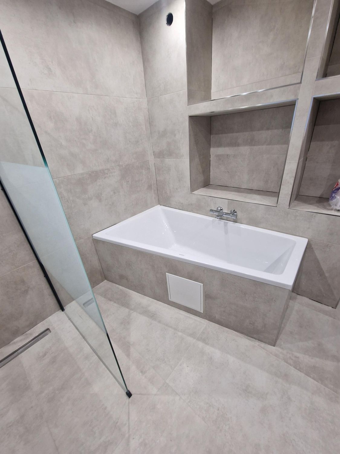 Salle de bains avec baignoire blanche, murs carrelés gris, porte de douche en verre et étagères intégrées.