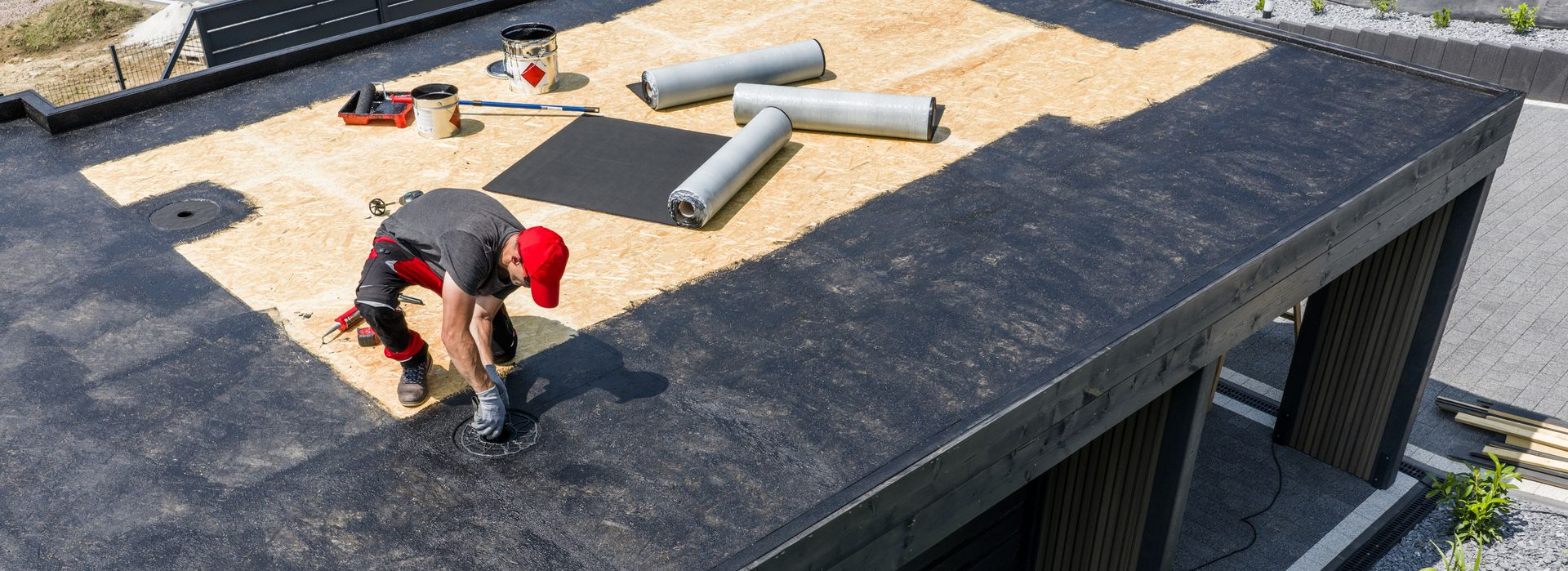 Un couvreur pose un revêtement de toiture noir sur un toit plat. Il porte un chapeau rouge et est agenouillé.