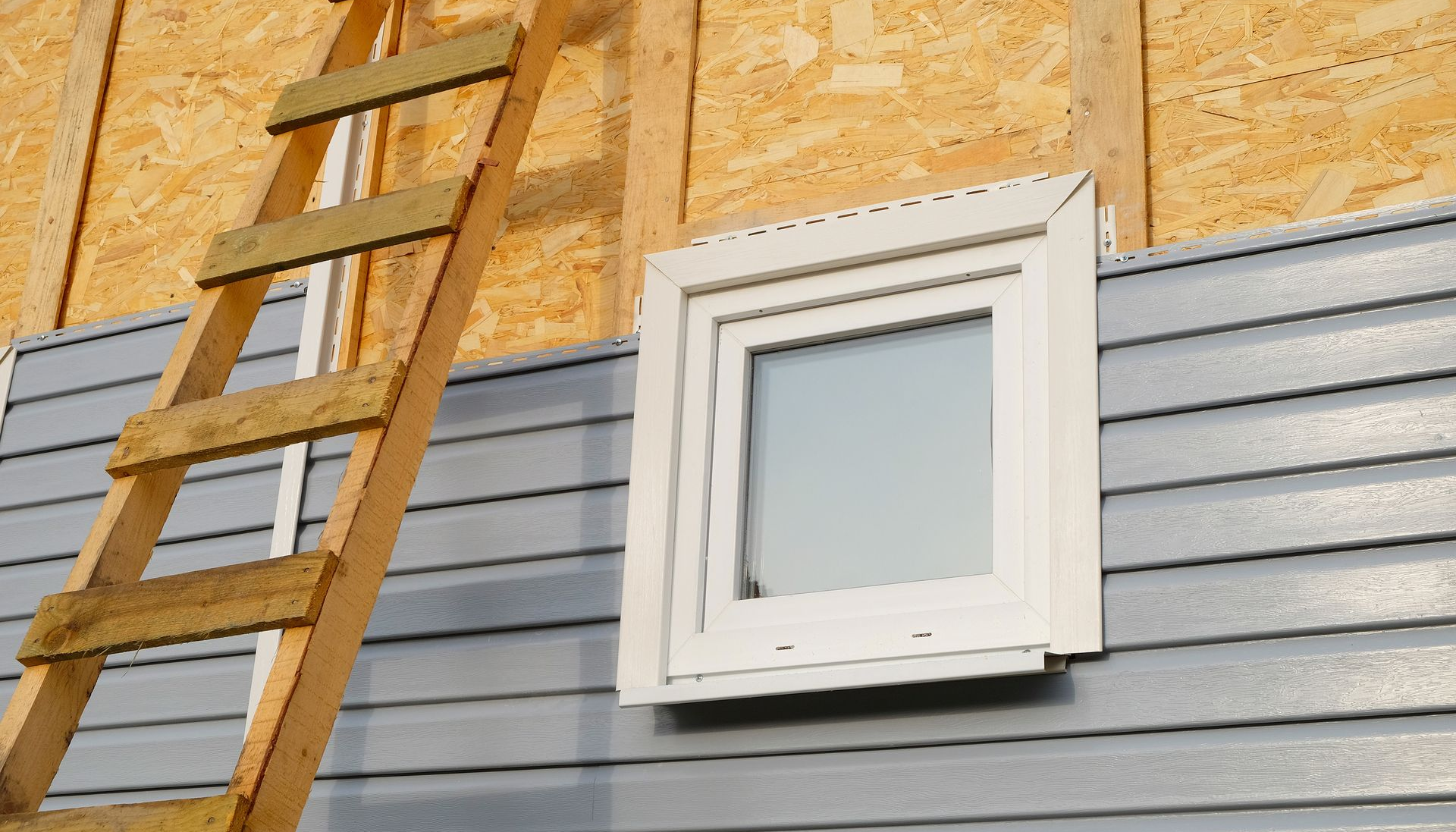 Échelle en bois à côté d'un bâtiment avec une fenêtre blanche et un bardage gris.