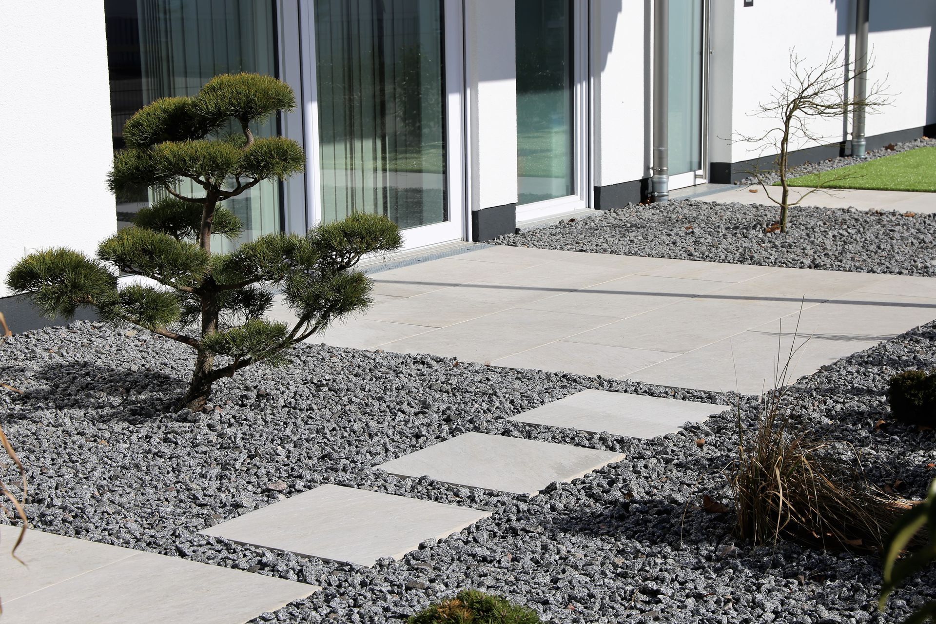 Des dalles carrées traversent un chemin de gravier gris, menant à un bâtiment moderne avec des portes vitrées et un aménagement paysager.