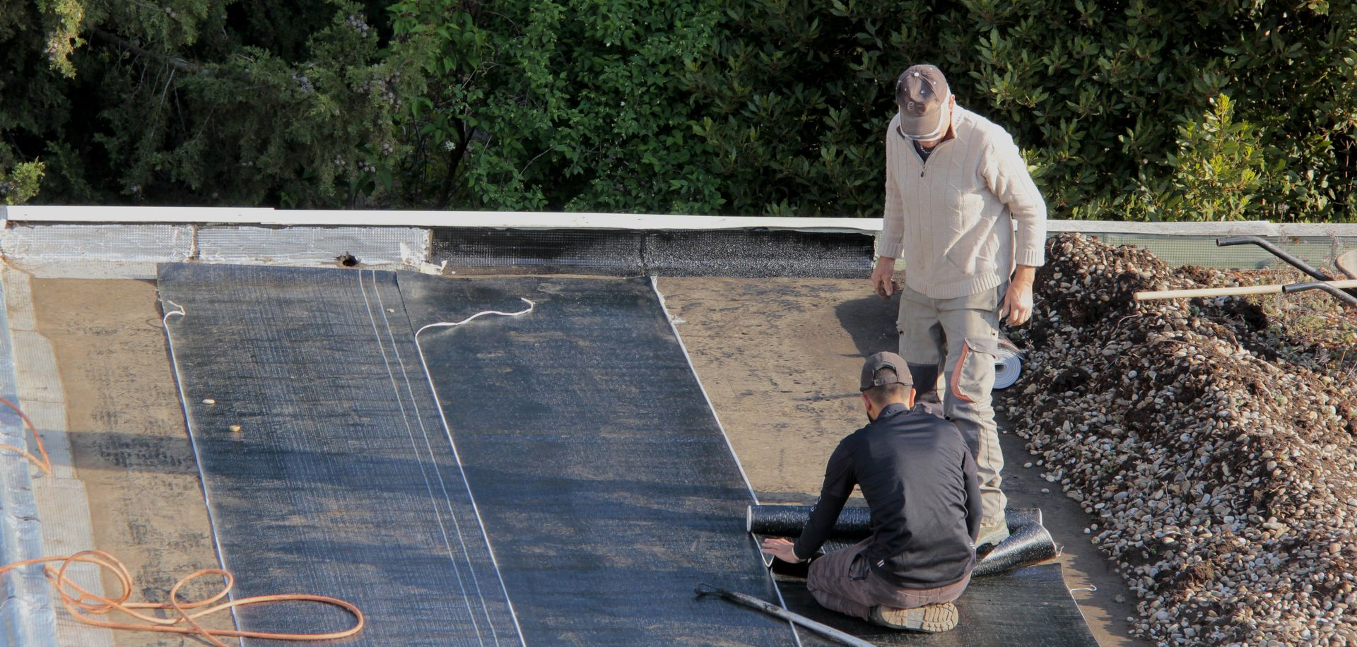 Deux hommes travaillent sur un toit plat, installant une couverture noire. Des arbres verts se dressent en arrière-plan.
