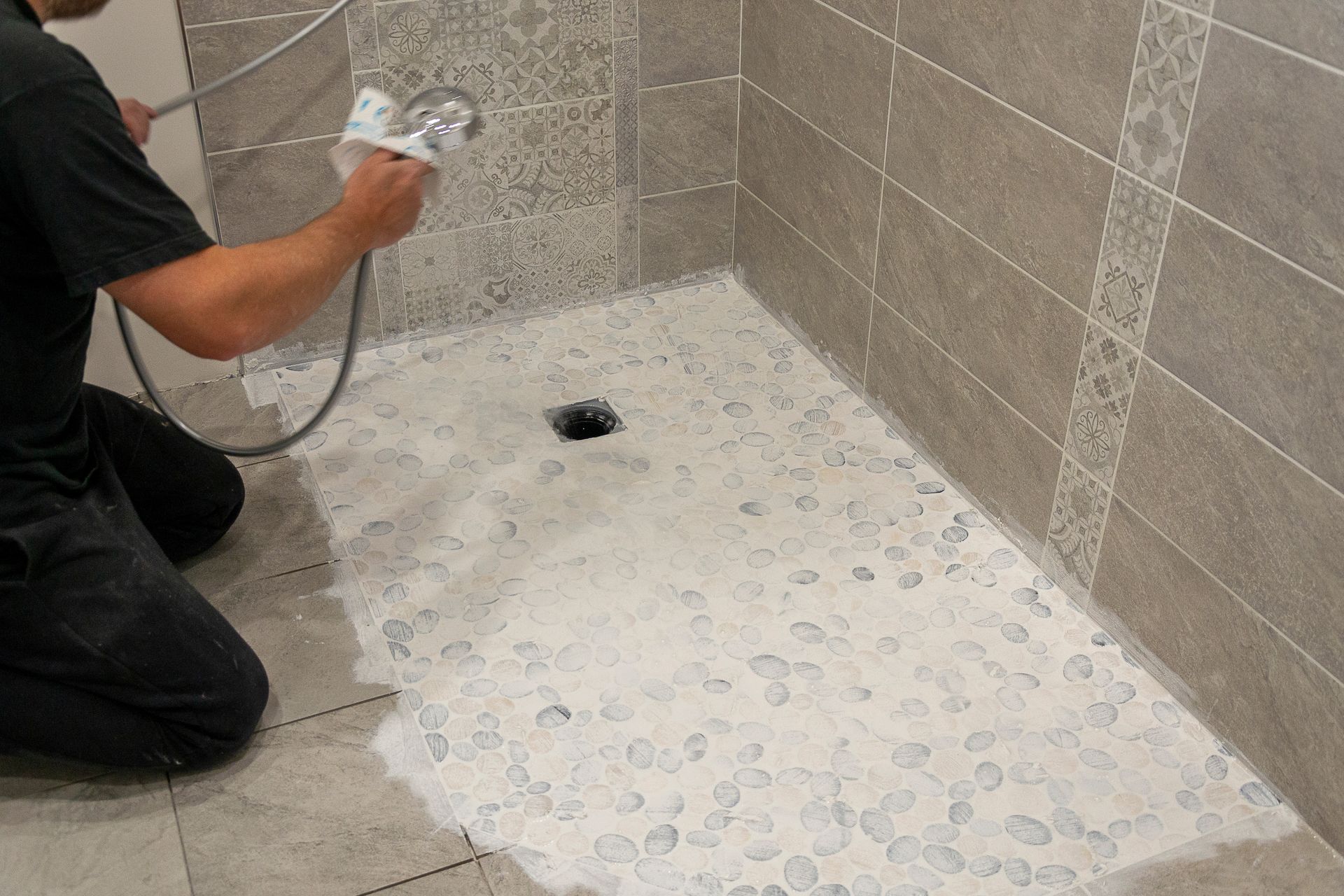 Personne effectuant les joints d'un carrelage en galets au sol d'une douche. Murs beiges, bonde noire, joints blancs.