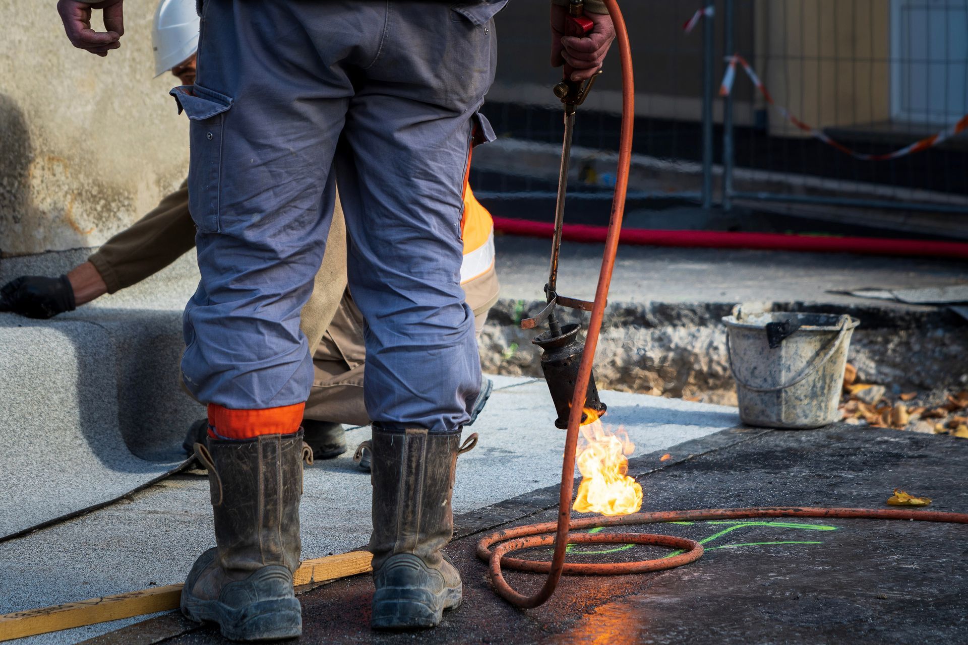 Un ouvrier utilise une torche sur la chaussée. Les flammes sont visibles. La personne porte des bottes de travail et un équipement de sécurité.