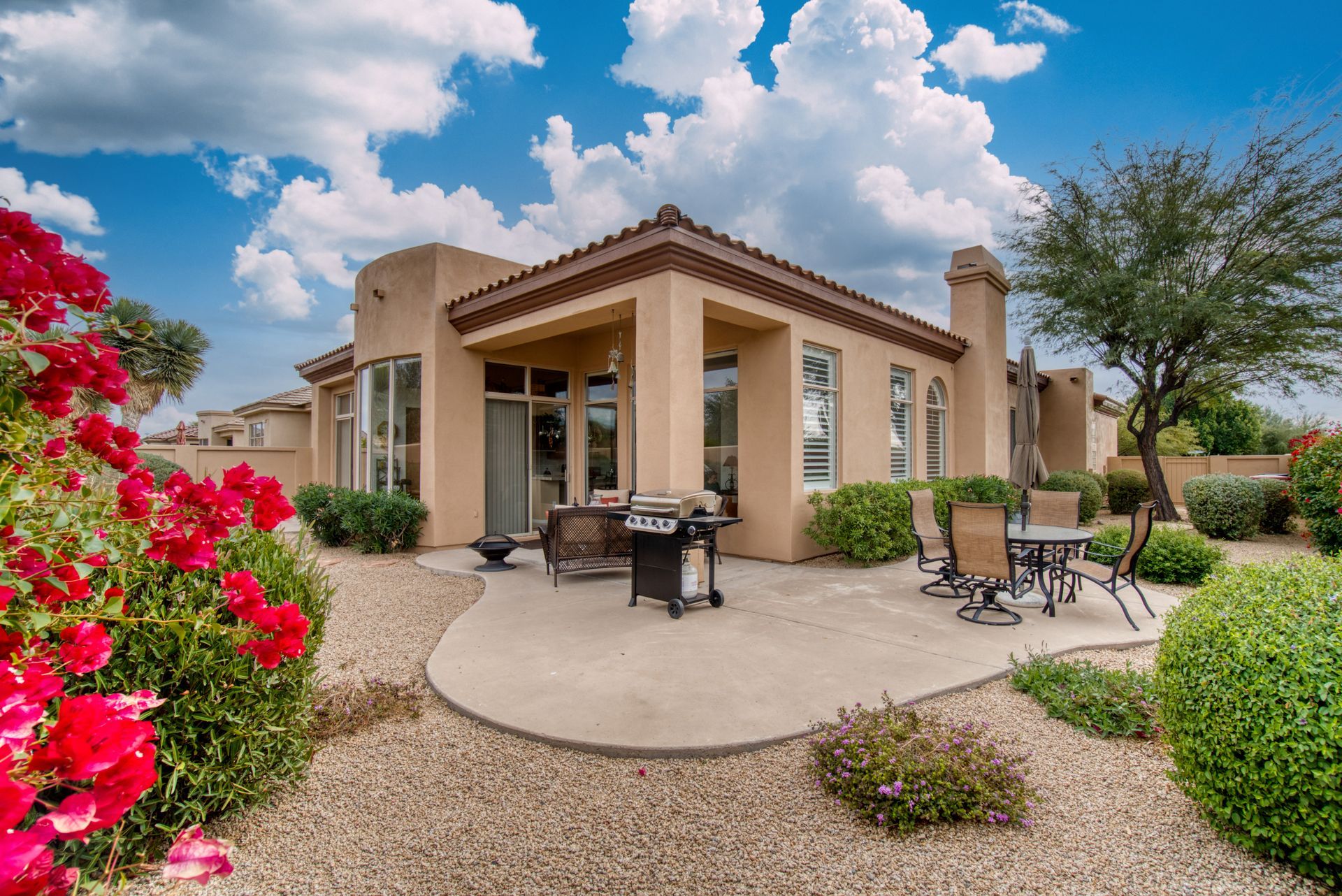 Terrasse arrière d'une maison en stuc beige avec aménagement paysager, ciel bleu et nuages.