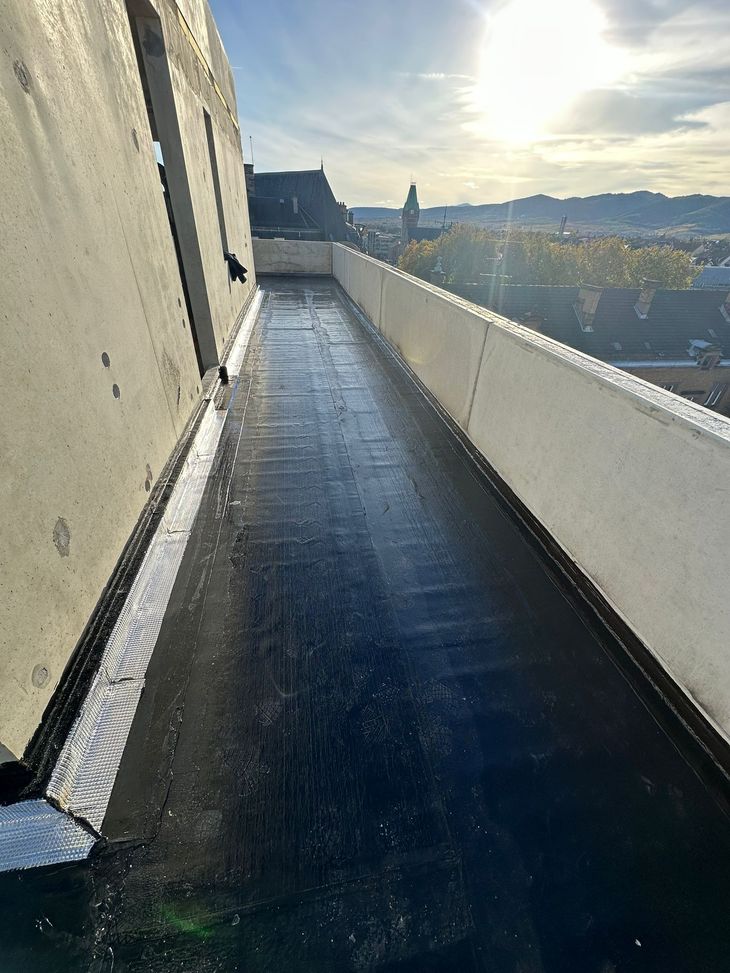 Toit-terrasse à la surface sombre et humide, bordé de murs blancs. Silhouette de la ville en arrière-plan, sous un soleil éclatant.