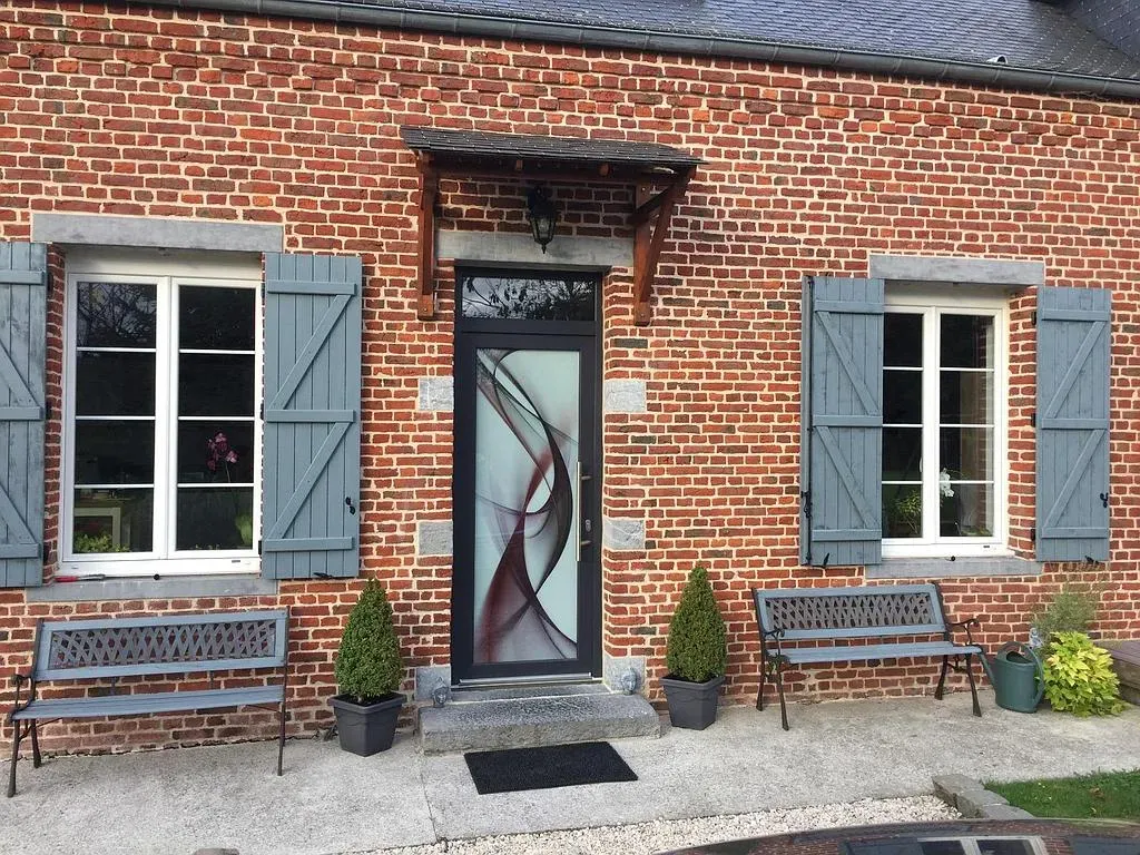 Façade en briques avec 2 fenêtres blanches à volets battants gris et une porte d'entrée noire avec motif sur la vitre