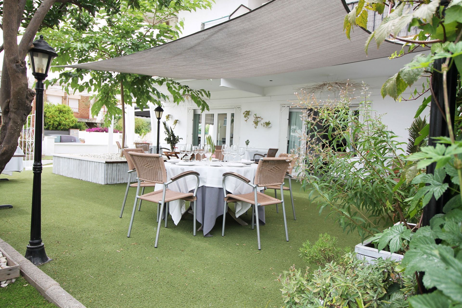 Comedor exterior en el patio con toldo blanco, sillas de mimbre y mesa redonda sobre césped verde rodeado de plantas.