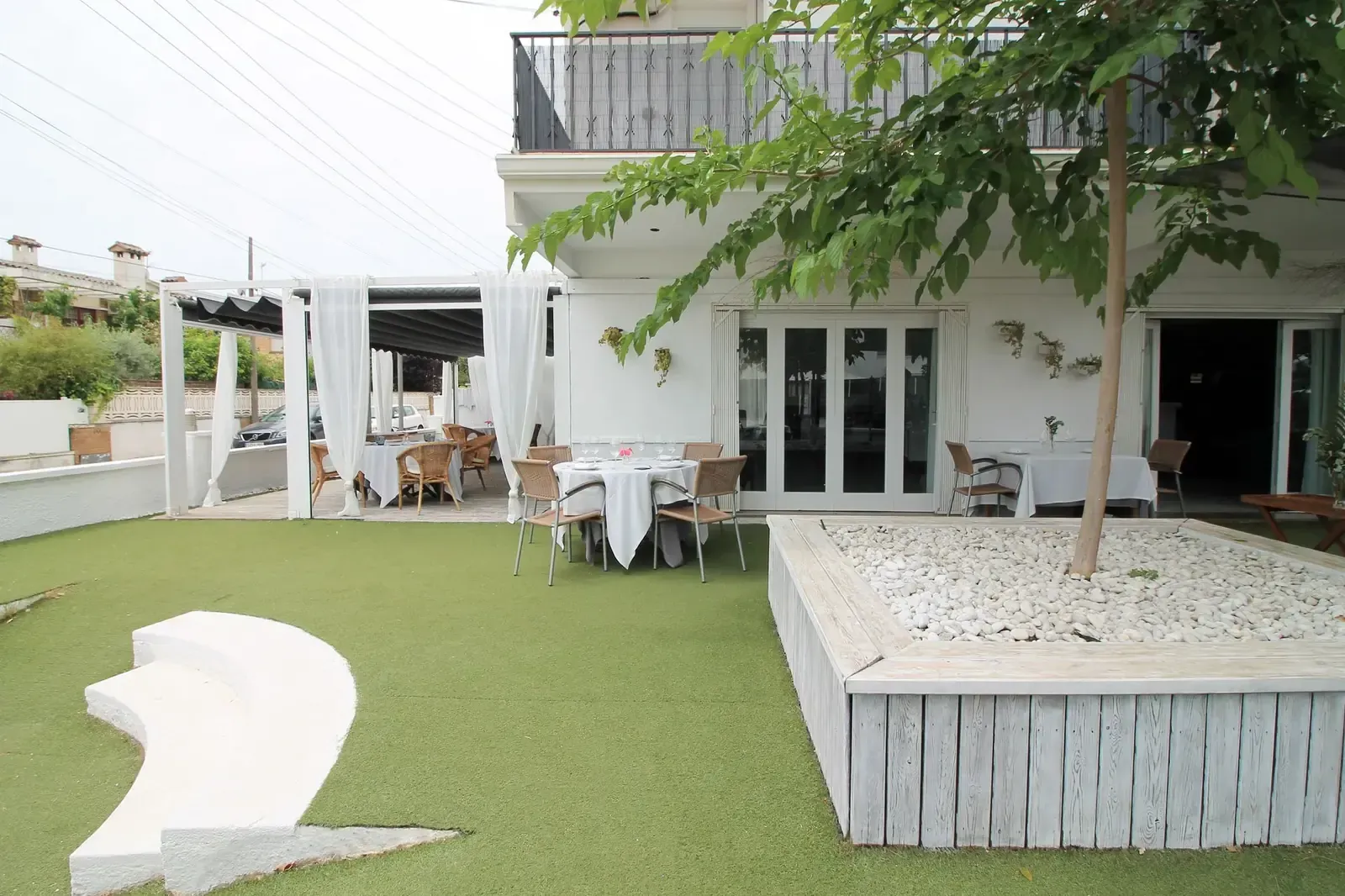 Patio moderno con edificio blanco, césped verde y asientos al aire libre bajo un árbol.