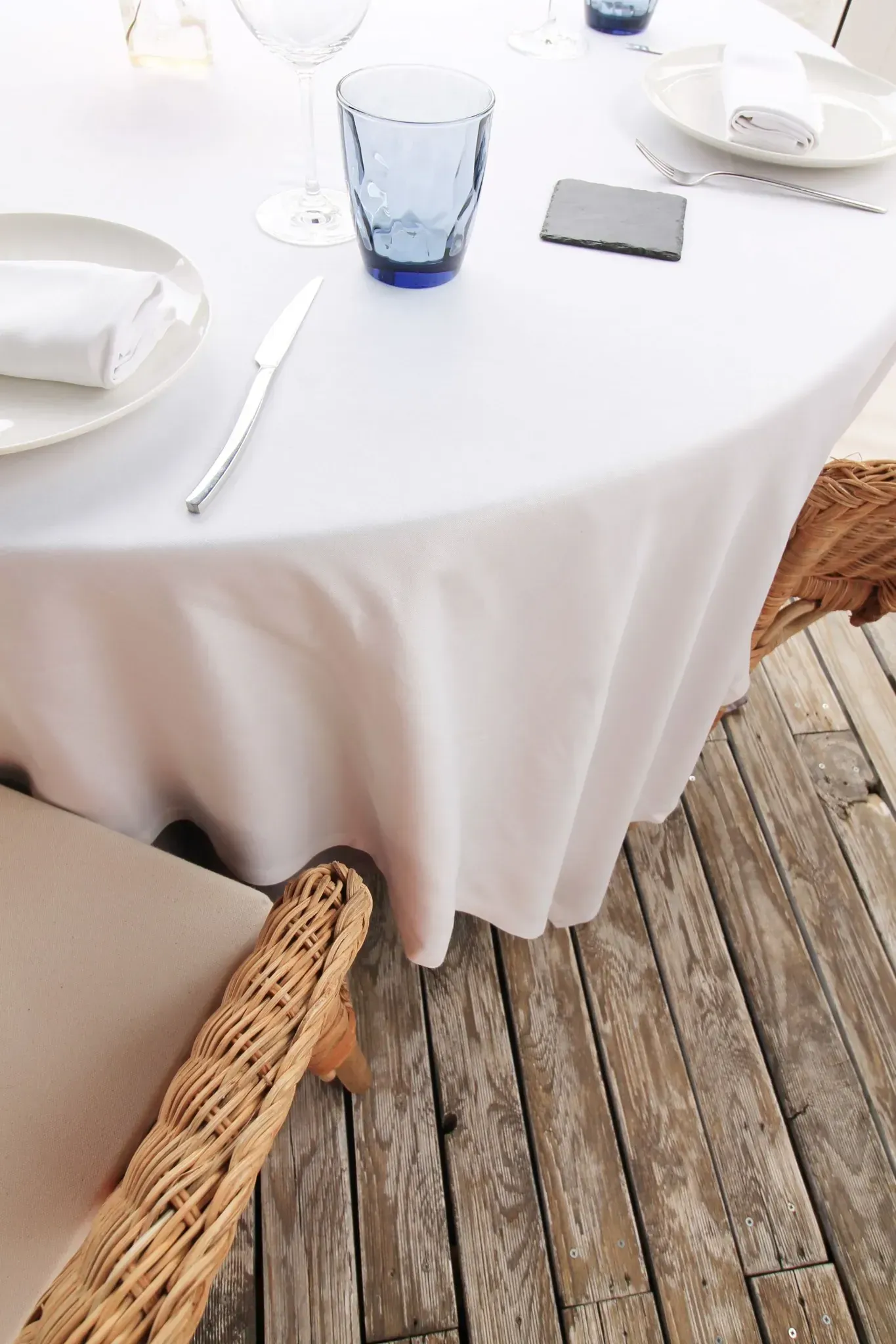 Mesa de comedor exterior cubierta con un mantel blanco, con vasos azules y sillas de mimbre sobre una terraza de madera.