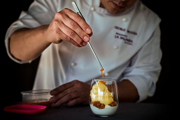 Chef rociando caramelo sobre un postre en un vaso sobre una mesa oscura.