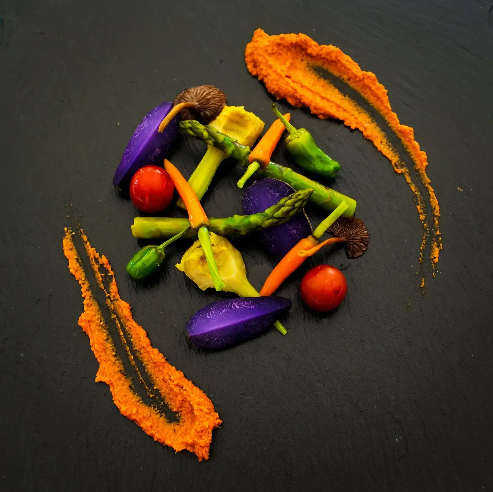 Verduras coloridas dispuestas artísticamente en un plato oscuro con vetas de salsa naranja.