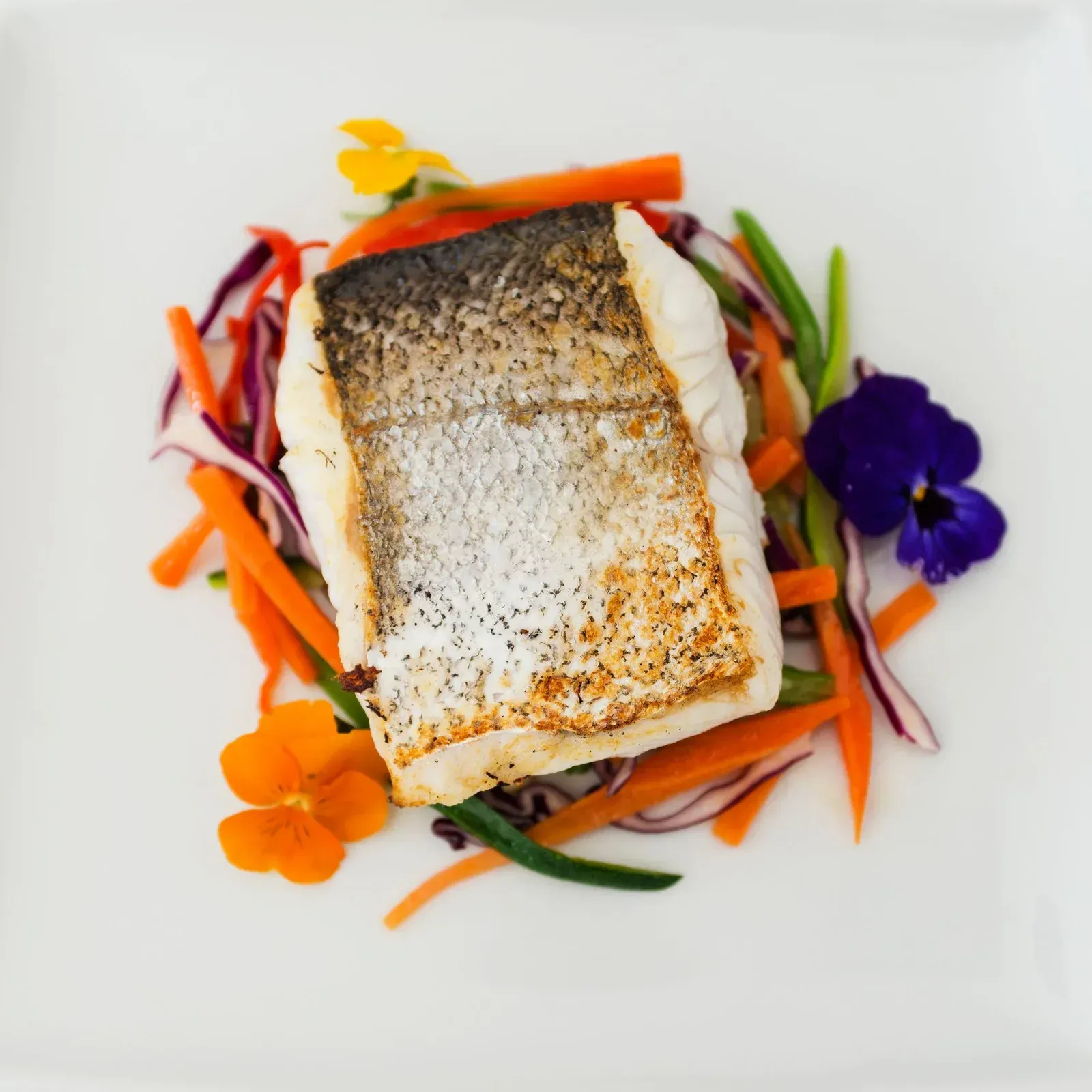 Filete de pescado sellado con coloridas cintas de verduras y flores comestibles en un plato blanco.